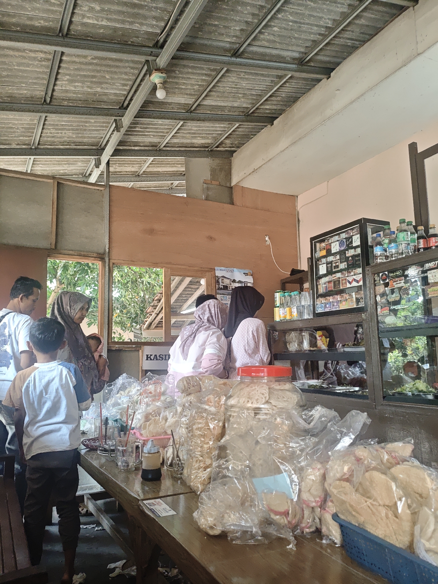 The image depicts an indoor scene at a shop in Lau, Indonesia, likely a small convenience store or warung. The environment appears rustic with a corrugated metal ceiling supported by metal beams, and wooden walls. Natural light filters in through two open windows in the background, revealing greenery outside.

Several people are present. On the left, a man in a white t-shirt and a young boy wearing a t-shirt with blue sleeves are standing. To their right, a woman in a brown hijab is holding a baby. Further to the right, two other women, both wearing hijabs and long-sleeved tops, are engaged in activity at a counter. One woman is wearing a light pink top with a polka dot pattern, and the other is wearing a darker pink top.

In the foreground, a wooden table is laden with various items. Several plastic-wrapped snacks or crackers are piled on the table, some in a blue basket. There are also glasses with red straws, a small bottle with a dark liquid, and other unpackaged snacks. Behind the table, a display cabinet filled with cigarette packs is visible, along with shelves stocked with bottled drinks and smaller jars. Another glass-fronted display case to the right contains more items, some appearing to be food. The overall impression is of a bustling, local shop environment.