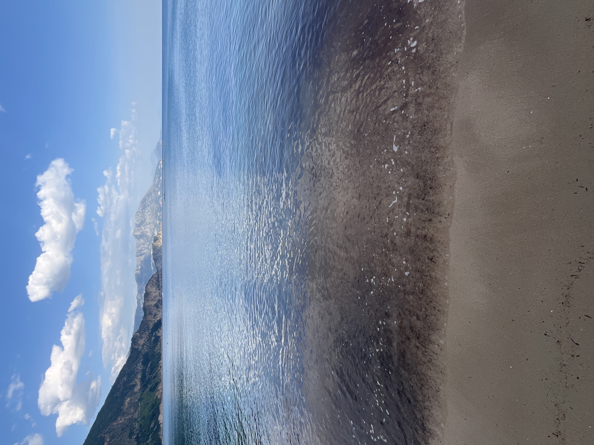 The image displays a serene coastal or lakeside landscape under a bright, clear blue sky with scattered white cumulus clouds. The perspective is from the water's edge, looking out across a body of water towards distant landmasses.
On the left side of the frame, when viewed upright, steep, dark-colored mountains or large hills rise directly from the water, extending into the midground. Patches of green vegetation are visible on their slopes. Further in the distance, additional layers of mountains are visible, their peaks appearing lighter and somewhat hazy against the sky.
The central and largest portion of the image is occupied by the calm water, which shimmers with reflections of sunlight, creating bright streaks on its surface. Small ripples are visible across the water's expanse. Closer to the shore, the water appears slightly darker and may contain some floating organic matter or debris.
The right side of the foreground features a strip of sandy or gravelly beach. The sand closest to the water is visibly wet and dark, while further inland, it is lighter and drier. Small specks of dark debris, possibly plant matter or small stones, are scattered across the beach.
No people or explicit activities are visible, emphasizing the natural beauty of the scene. The weather is clear and sunny, indicative of daytime, likely morning or early afternoon. The mountainous coastline and clear water are consistent with the location context of Prijevor, Montenegro. No visible text is present in the image.