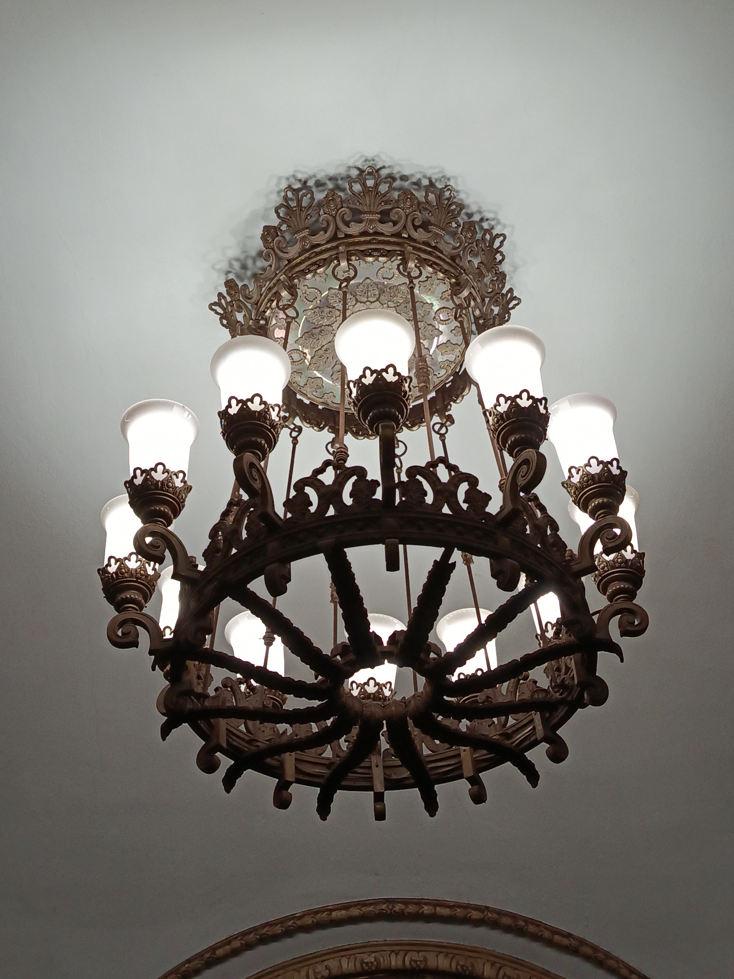 A low-angle shot shows a vintage chandelier hanging from a white ceiling. The chandelier has a circular bronze frame decorated with ornate carvings. Ten frosted glass lamps are arranged around the circular frame, emitting a soft glow. The chandelier is suspended from an equally ornate bronze fixture attached to the ceiling. This architectural detail can be found in the Kiyevskaya station in the Moscow Metro system, Moscow, Russia. A partial view of a similar decorative molding is visible in the lower foreground.