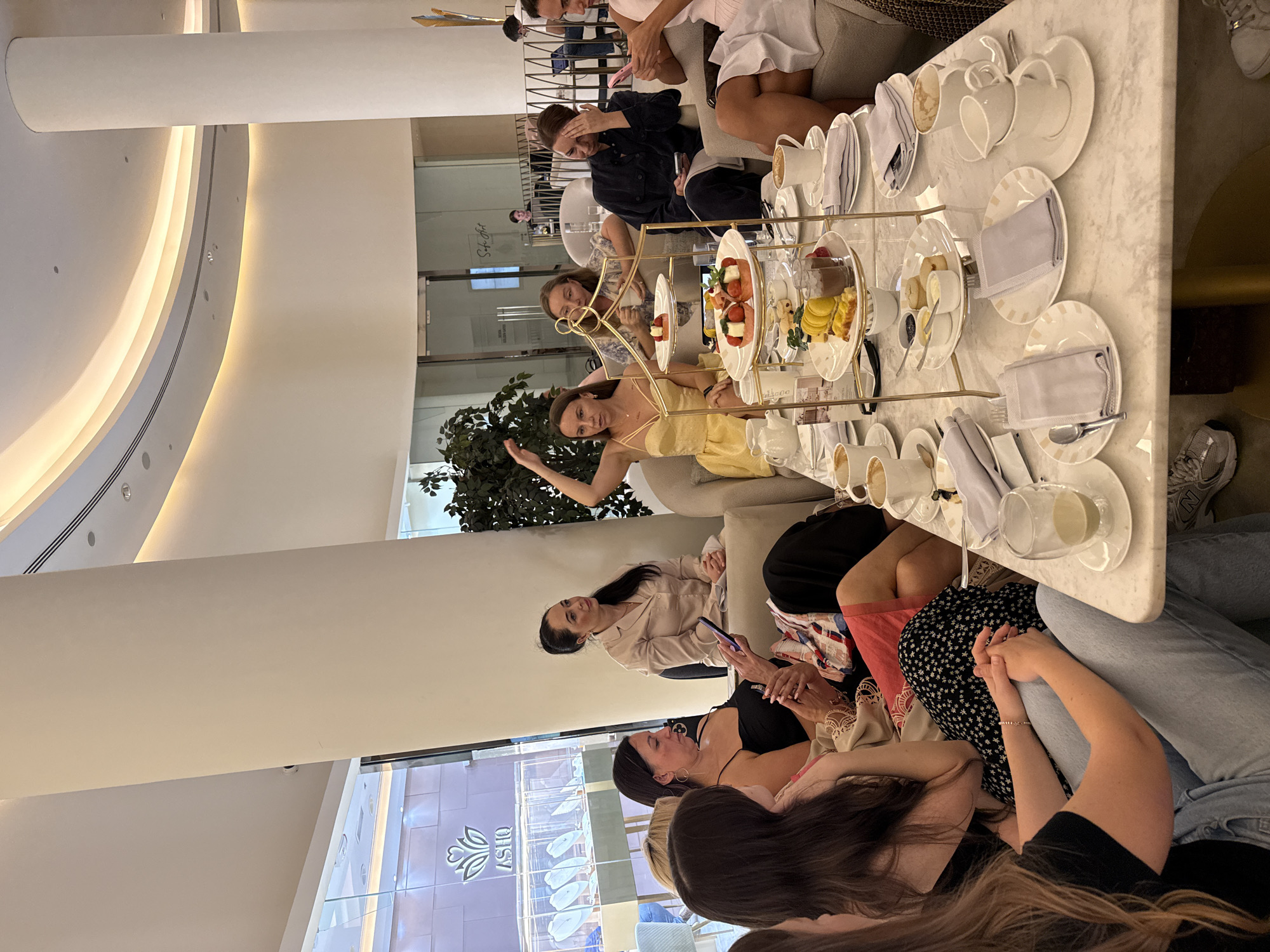 An upscale indoor setting, likely a hotel lounge or high-end cafe in Dubai, is depicted during what appears to be a high tea or afternoon tea gathering. The spacious venue features modern, clean architecture with curved white walls, large columns, and bright, indirect lighting from recessed fixtures.
Numerous women are the primary subjects, seated in groups on light-colored couches and chairs around marble-topped tables. They appear to be engaged in social interaction, conversation, and enjoying the refreshments. One woman in a yellow dress is seen leaning back on a sofa, gesturing with her left hand, while others are seen talking or looking at their phones.
Each table is elegantly set with white porcelain teacups, saucers, small pitchers, and plates with folded gray napkins. Prominently featured are multi-tiered high tea stands laden with a variety of pastries, cakes, scones, small sandwiches, and fresh fruits like strawberries and sliced mandarins. Empty and partially used cups suggest the event is in full swing.
In the background, through glass partitions, other areas of the establishment are visible, including what appears to be a retail space with the name "ASHQ" and a floral logo prominently displayed. Another glass door partially shows text that might read "Safina Art" or similar. The overall ambiance is relaxed and sophisticated, typical of a luxurious social gathering. The bright lighting suggests it is daytime.