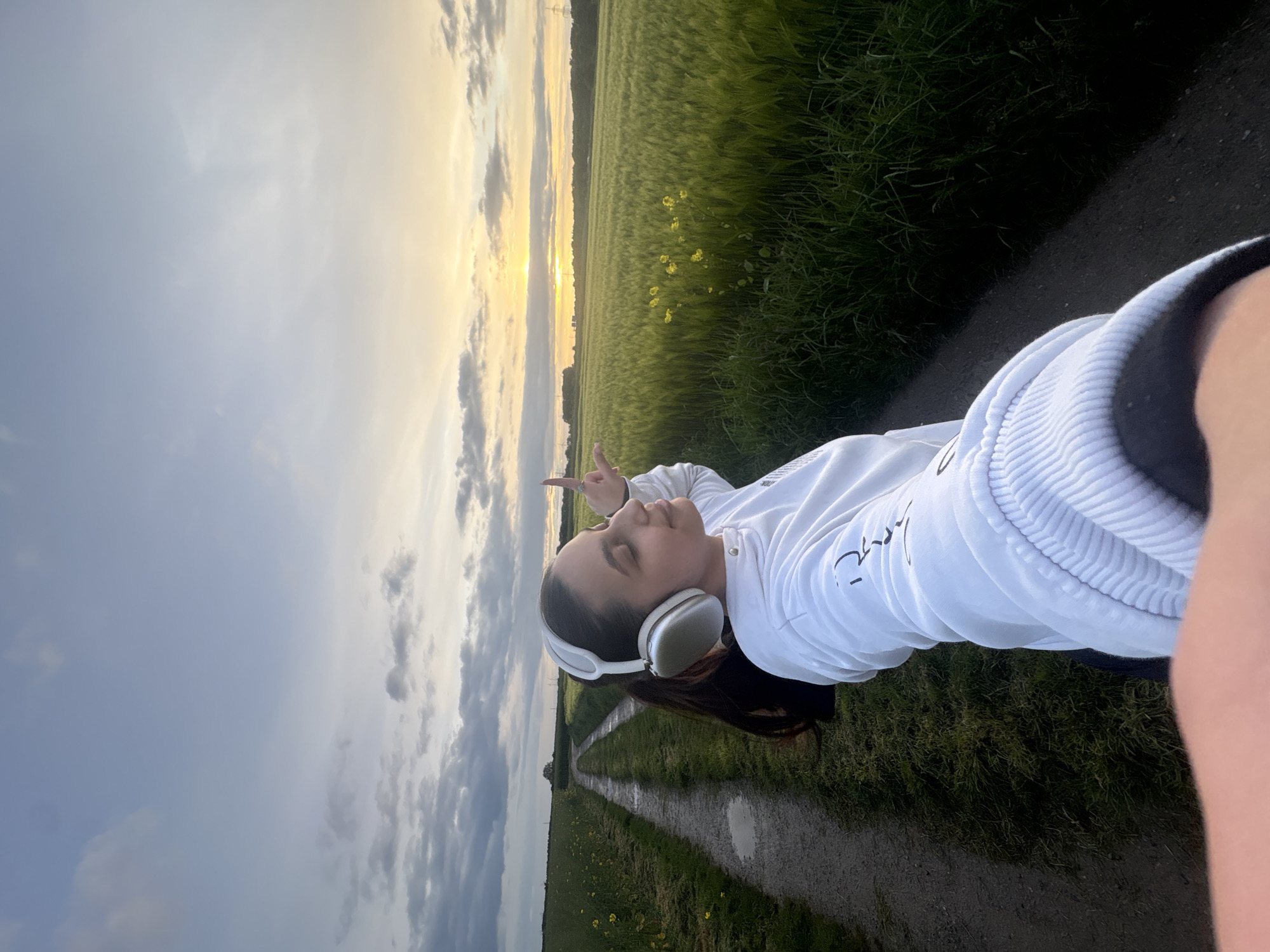 The image depicts a person in a white sweatshirt and headphones standing on a path in a field at dusk or dawn. The field is likely in Mönchengladbach, Germany. The person is making a peace sign with their hand. The environment consists of a dirt or gravel path running through a field of tall, green grass or wheat. There are scattered yellow flowers. The sky is partly cloudy with the sun near the horizon. The orientation suggests a selfie.