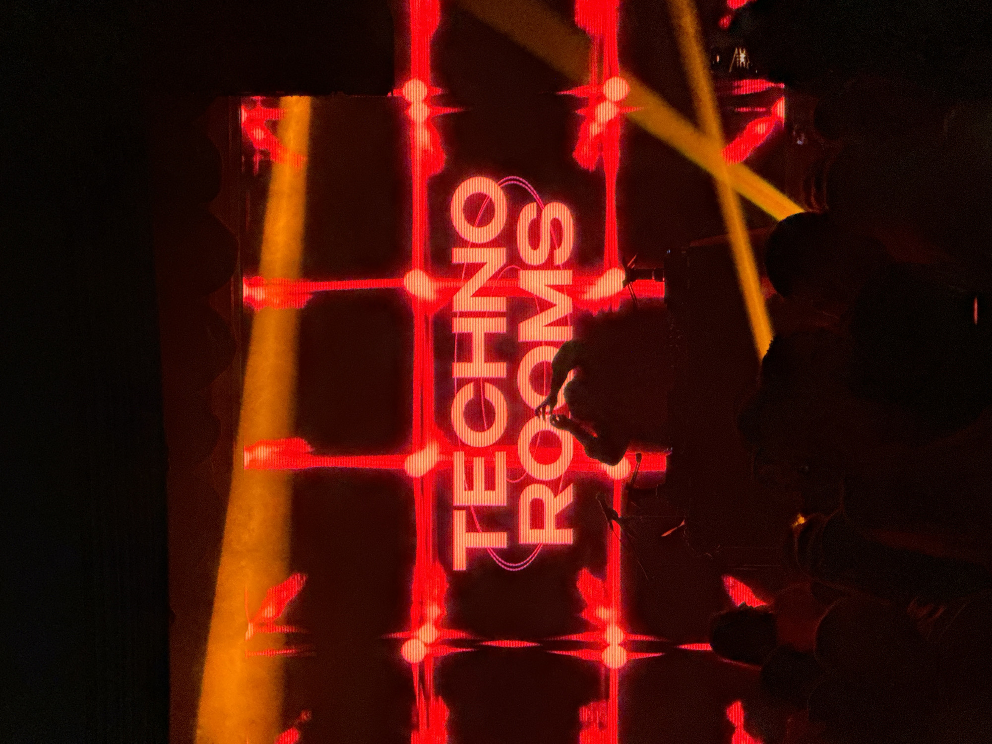 The image shows a person standing in front of a screen displaying the words "TECHNO ROOMS" in red neon lights. The background consists of a grid-like structure also illuminated in red. There is a vertical beam of yellow light on the left side of the screen and a diagonal yellow beam on the right. The person is partially obscured by shadows, but their face is illuminated by the screen's light. The scene appears to be indoors, possibly at a club or event venue. The location is Moscow, Russia.