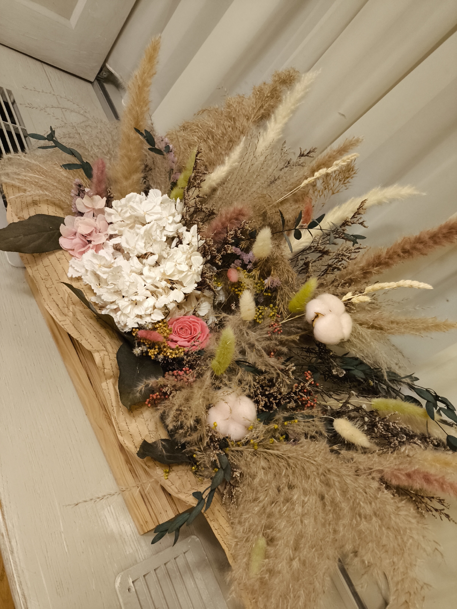 The image shows a dried flower arrangement. The arrangement features white hydrangeas, pink roses, cotton, and pampas grass in shades of brown, cream, and pink. The arrangement is wrapped in paper. The setting appears to be indoors, with a white paneled wall in the background. The location is not Smolensk, Russia.