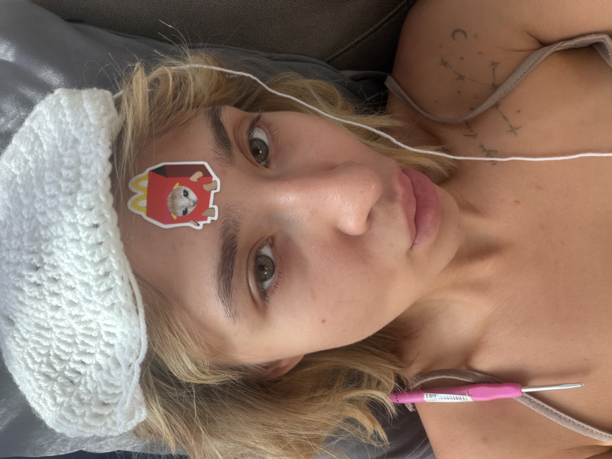 This is a close-up, over-the-shoulder shot of a young person lying down, their face turned towards the camera. They have blonde hair and light-colored eyes, with a sticker of a cat in a McDonald's box on their forehead. A white crochet headband is on their head, and white yarn runs across their face and chest. A pink crochet hook is visible on their collarbone. The person is wearing a light-colored tank top. There is a tattoo on their shoulder with astrological symbols, including a crescent moon and stars. The person's expression is neutral, and their tongue is slightly sticking out. The background appears to be a gray sofa. The lighting suggests it is daytime.