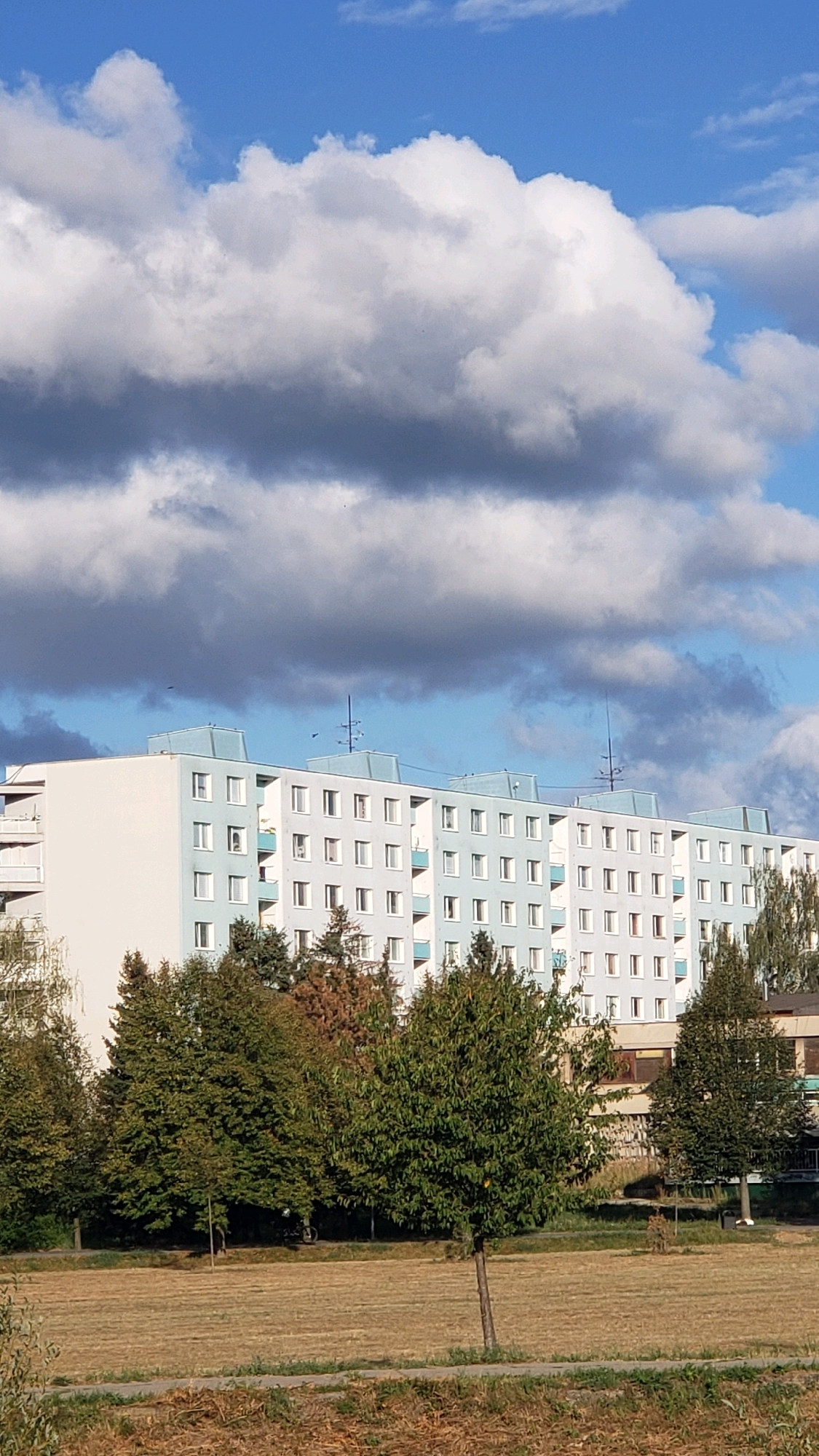 The image depicts a pale blue and white apartment building, likely in the city of Prešov, Slovakia, under a cloudy sky. The building features multiple stories with uniformly spaced windows and balconies. In front of the building, a line of trees, some with green foliage and others with autumnal colors, separates it from a dry, mowed field. A few small trees are scattered within the field. The sky is a mix of bright blue and heavy grey clouds, suggesting dynamic weather. There are no people visible in the scene.