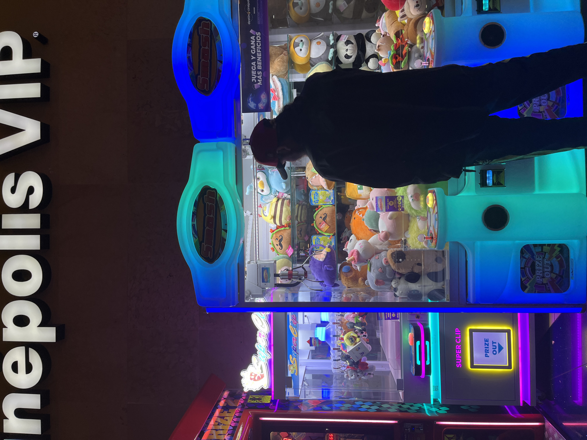 A person is standing in front of a claw machine at Cinépolis VIP in Puebla, Mexico. The claw machine is brightly lit with blue, green, and pink neon lights. The top of the machine is filled with plush toys. The lower machine is labeled "SUPER CLIP" and has a "PRIZE OUT" sign illuminated in yellow. The person is wearing a dark jacket and a red hat and appears to be focused on the plush toys within the machine. A sign reading "Cinepolis VIP" is visible on the left side of the frame.