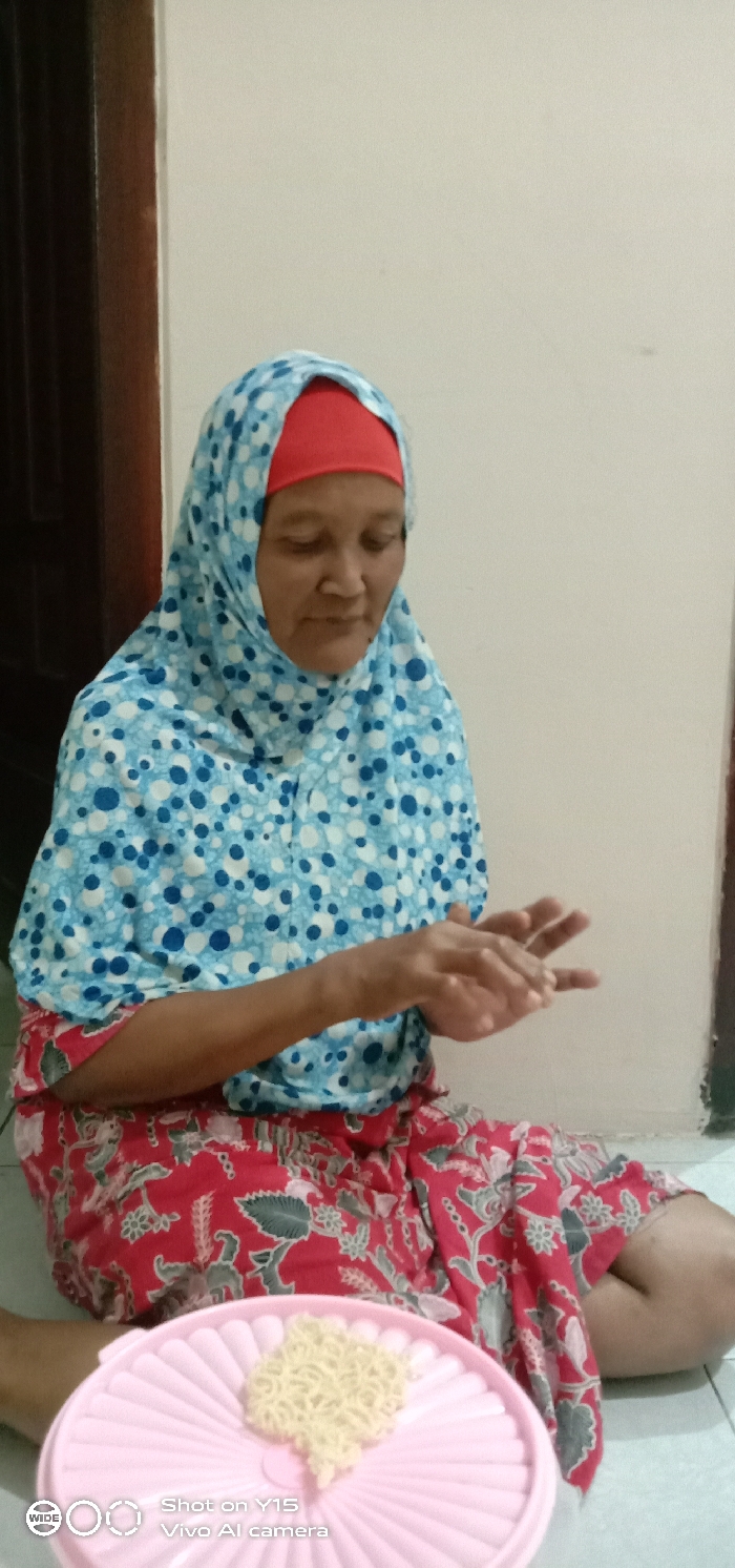 In Ranugedang, Indonesia, an adult female is seated on the floor in an indoor setting. She wears a light blue hijab adorned with white and dark blue polka dots, covering a red headwrap. Her lower body is clad in a red patterned garment, possibly a sarong, featuring grey or greenish floral and foliage designs. Her hands are raised and held in a partially cupped position in front of her upper body, with her gaze directed downwards. In the foreground, positioned on a light-colored floor, is a light pink plastic container lid, ridged with a fan-like pattern. On this lid rests a small, heart-shaped cluster of light yellow, curly uncooked instant noodles. The background consists of a plain, light-colored wall, with a dark brown doorframe visible on the left side of the frame.