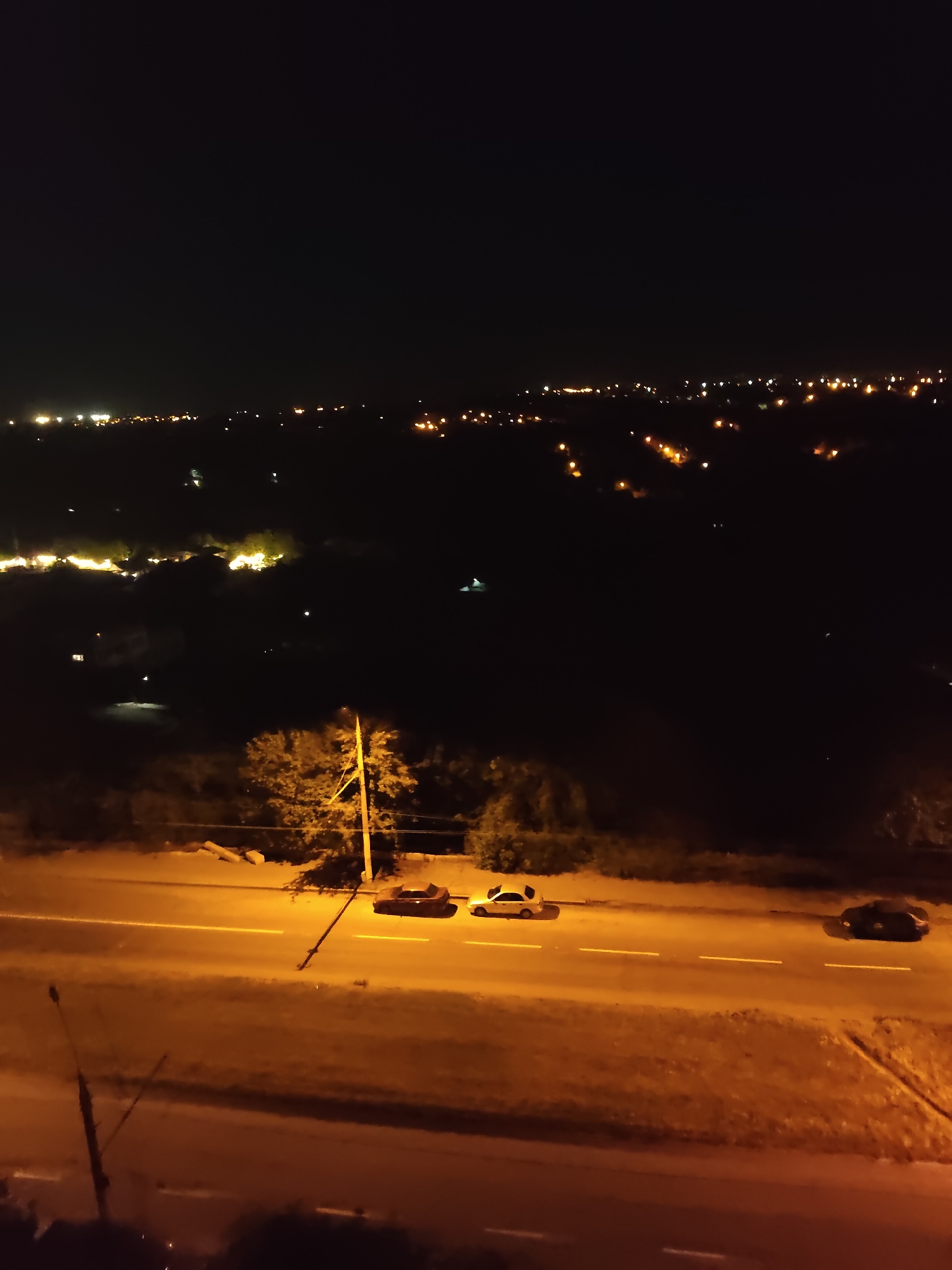 This is a nighttime, high-angle, full shot of a highway in the city of Zaporizhzhia, Ukraine. The highway is illuminated by streetlights, casting a warm, orange glow on the asphalt and surrounding vegetation. Three cars are visible on the highway. Two cars are parked side-by-side in the left lane, and a third car is traveling in the right lane. In the background, a city skyline with numerous lights can be seen stretching across the horizon. A single utility pole stands near the edge of the highway, with wires extending from it. The overall scene is one of a quiet, urban night.