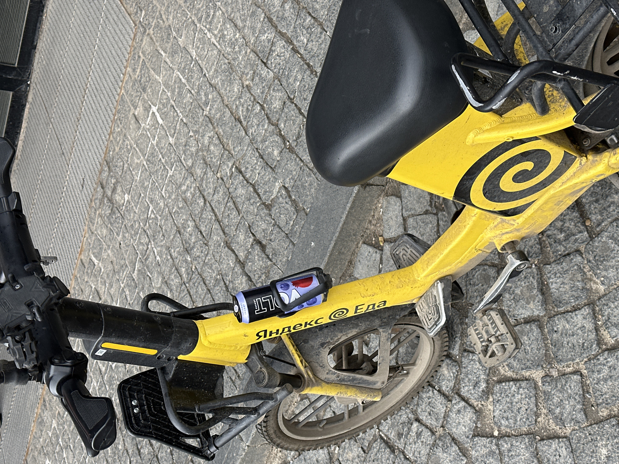 A yellow Yandex Eda bicycle is parked on a cobblestone street in Nizhny Novgorod, Russia. The bicycle has a black seat and handlebars. A can of Bolt beverage is attached to the bike frame. The bicycle is positioned at an angle, with parts of the front and back visible. The cobblestone pavement is textured.