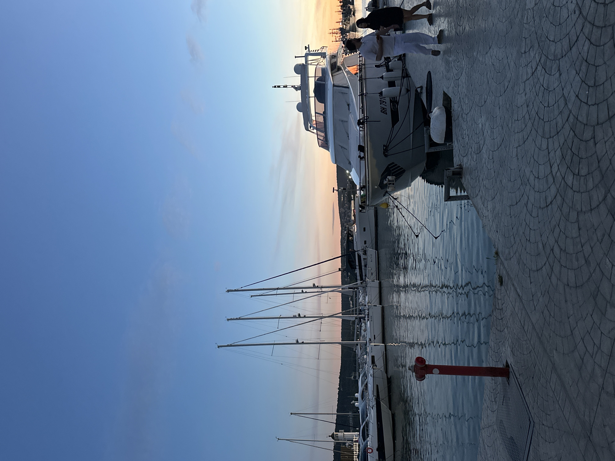 The image depicts a marina at sunset. A white motor yacht with the registration number "BH 7910" is docked on the right side of the frame, and two people are visible on the deck. Several sailboats with tall masts line the marina in the background. A red fire hydrant stands on the edge of the paved dock near the water. The sky is a gradient of blue to orange, indicating the time of day. The water reflects the colors of the sky and the docked boats. The marina is located in Varna, Bulgaria.