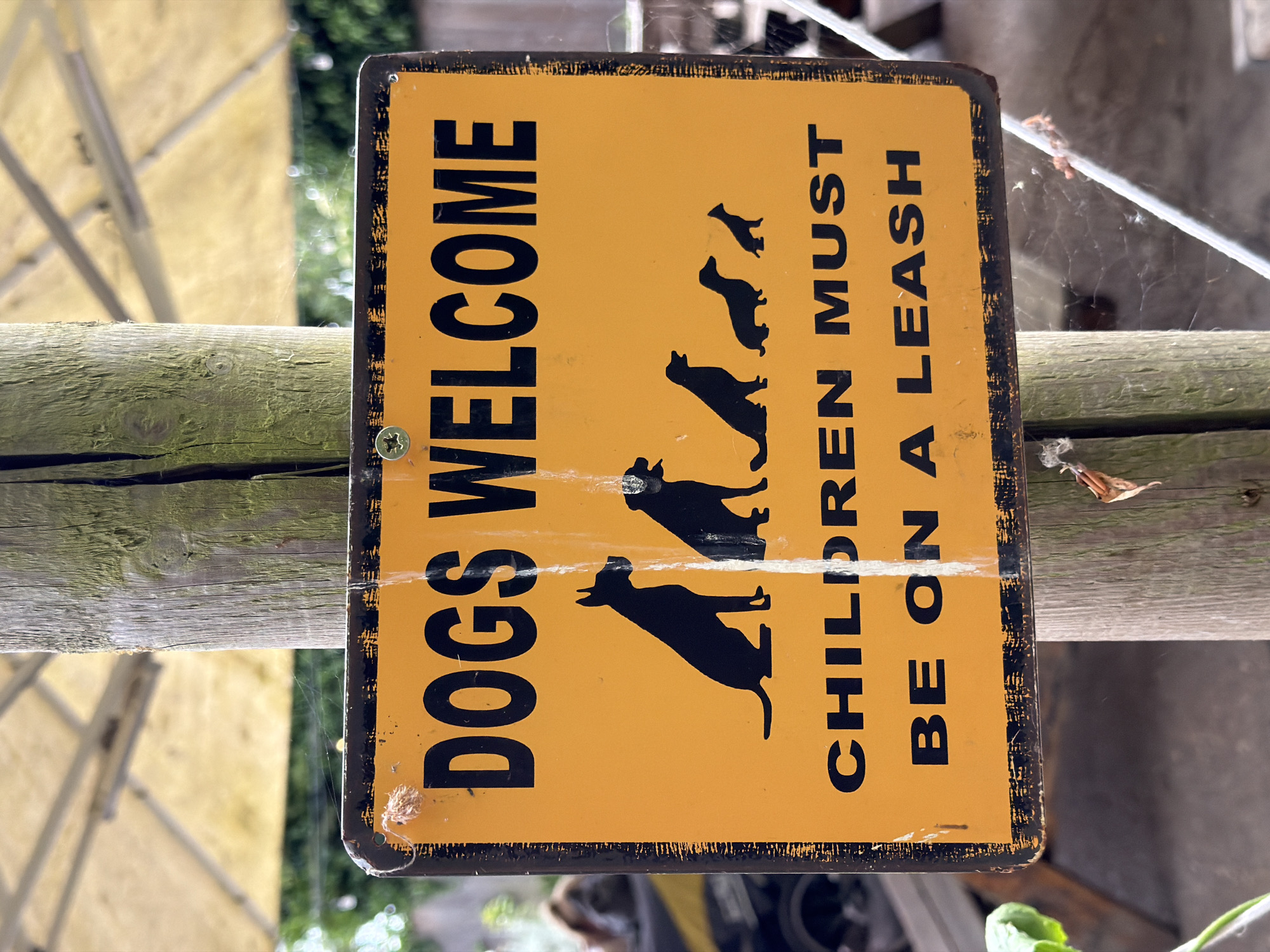 The image shows a yellow and black sign with the words "DOGS WELCOME CHILDREN MUST BE ON A LEASH". The sign is attached to a wooden post. The background includes blurred foliage and a glimpse of what might be an umbrella. The location appears to be an outdoor area, possibly a park or garden. The specific location within Folkestone and Hythe District, United Kingdom, cannot be determined from the image.