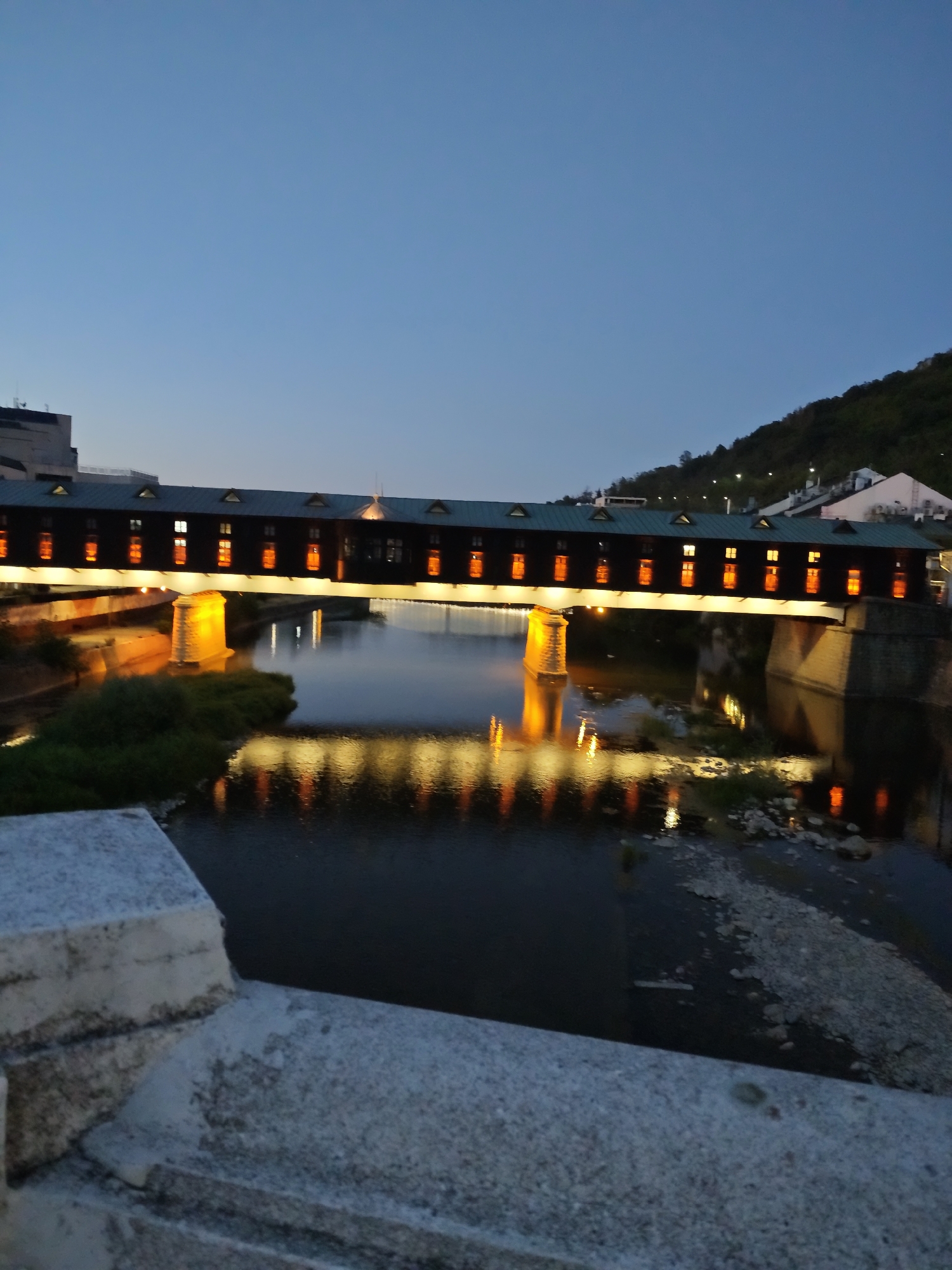 The image captures the iconic covered bridge of Lovech, Bulgaria, at dusk. The bridge, illuminated by warm light emanating from its numerous windows and supporting structures, spans a calm river. The reflection of the bridge's lights shimmers on the water's surface, creating a symmetrical and serene scene. The sky above is a deep blue, transitioning from twilight to night. The riverbanks are lined with vegetation, and in the background, a hillside dotted with houses and streetlights slopes upwards. The foreground features a stone parapet, part of a viewing platform or another bridge, providing a grounded perspective on the architectural marvel. The image is a static, wide shot, with no people or animals visible, focusing solely on the interplay of light, water, and the historic bridge.