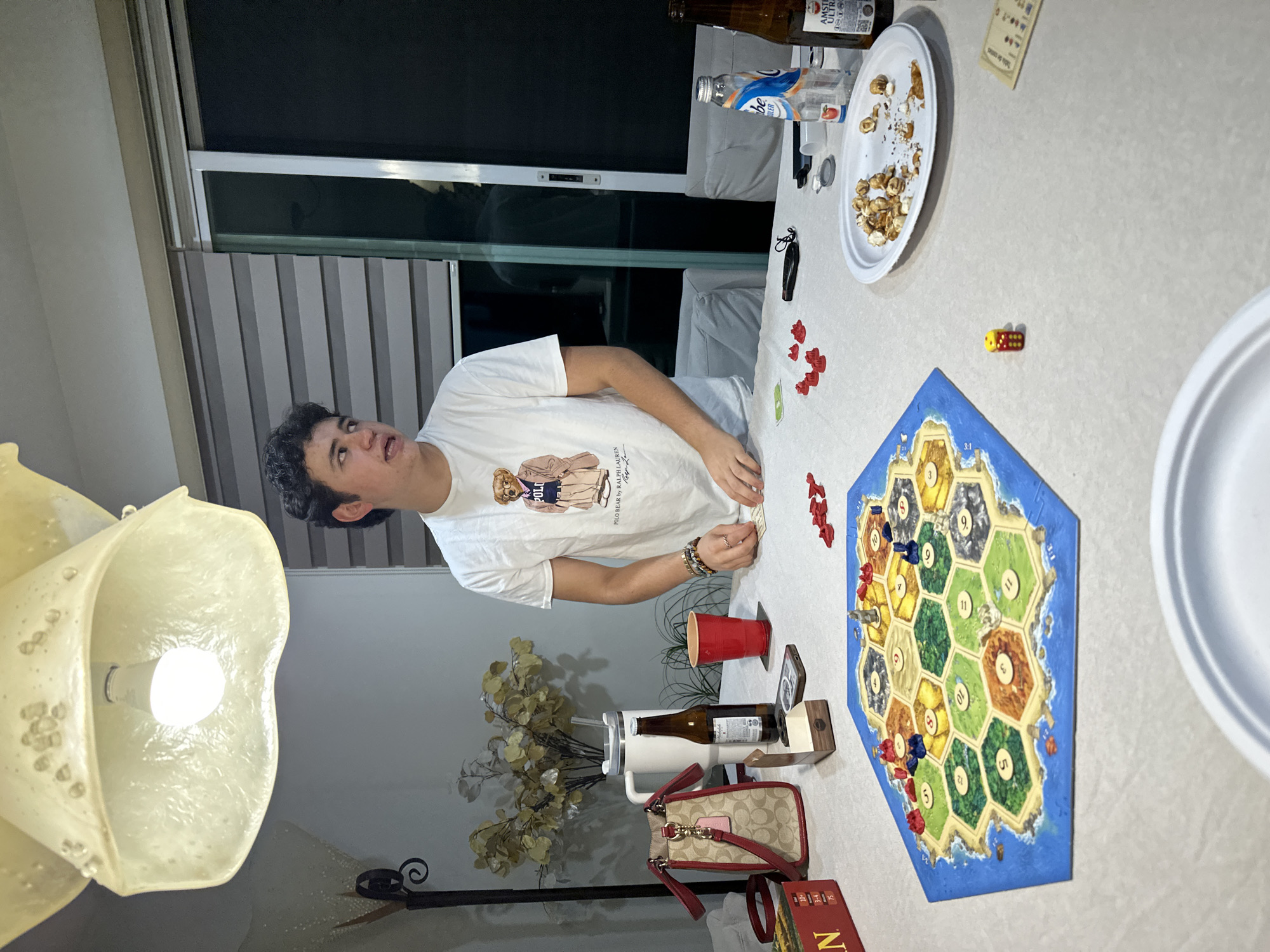 A young man with curly dark hair, wearing a white t-shirt with a graphic of a teddy bear in a preppy outfit, stands at a table in what appears to be a dining room or living area. He is looking to his right, with his mouth slightly open as if speaking. In front of him, a Catan board game is set up with hexagonal tiles depicting various terrains and numbers. Red and blue game pieces are scattered on the board. A yellow die with red dots sits on the table near the game.
The table is covered with a white tablecloth and is laden with several items. These include a red plastic cup, a brown bottle of beer, a white insulated tumbler with a straw, a small Coach handbag with red trim, a wooden game piece holder, a smartphone, and a white paper plate with popcorn and scattered crumbs. To the right of the plate, there is another bottle of beer and a clear plastic bottle with a blue and white label. In the background, a sliding glass door with blinds is visible, suggesting an indoor setting at night. The overall atmosphere appears casual, with a group of people engaged in a board game, likely a social gathering. The lighting is warm, coming from a decorative ceiling light fixture above the table. The scene is consistent with a domestic setting, possibly in a home in San Bernardino Tlaxcalancingo, Mexico, given the provided location.