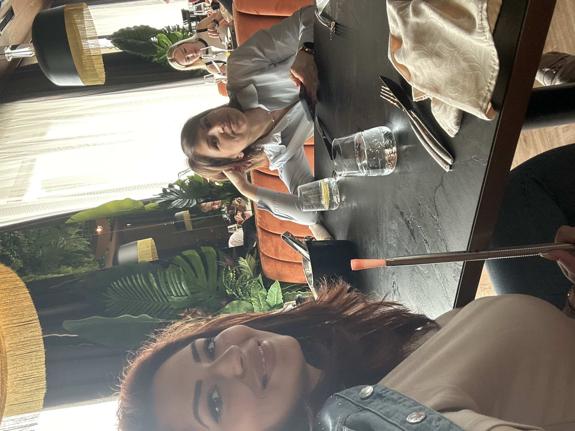 The image captures an indoor scene in what appears to be a restaurant. Three women are visible, seated at a dark-colored table. One woman is centered, leaning on her hand and looking towards the camera. Another is slightly off to the side, visible behind the first woman. The third woman is in the foreground, facing toward the camera, holding a hookah pipe. On the table, there are glasses with water, cutlery, and a light-colored napkin. Lush green plants and decorative lamps are visible in the background. This image does not have enough information to determine the exact location in Sofia, Bulgaria.