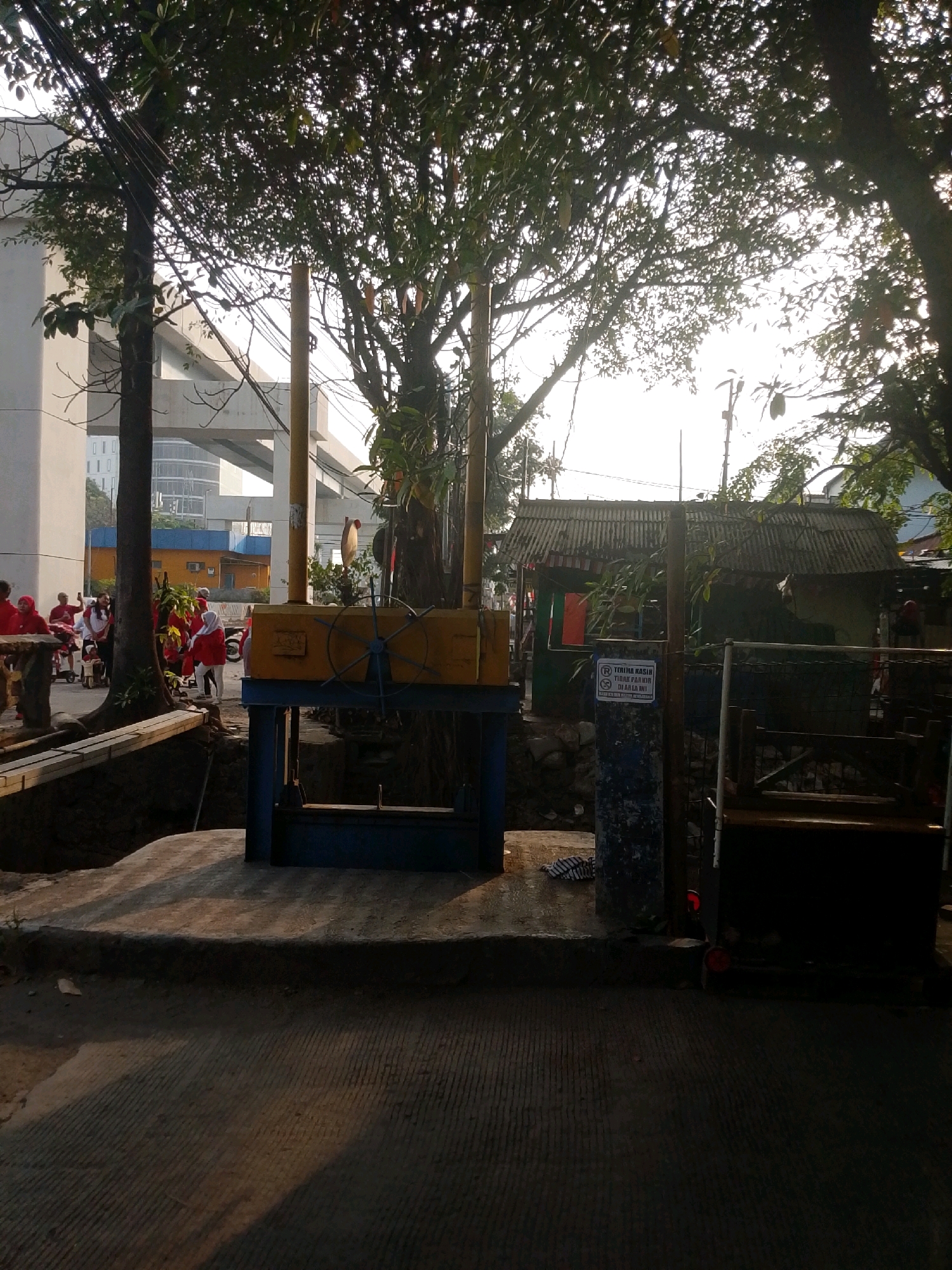 In East Jakarta, Indonesia, on a street with a paved surface, a water pump mechanism is visible. The mechanism is blue and yellow, with a wheel and metal parts. A sign with Indonesian text, including "TERIMA KASIH" and "DILARANG PARKIR DI AREA INI," is posted on a blue pillar. In the background, trees with dense foliage cast shadows, and a corrugated metal roof structure is present. A concrete overpass is also visible. Several people in red clothing are gathered near a wooden bench on the left side of the image, suggesting a community event or gathering. The scene is illuminated by natural light, possibly from early morning or late afternoon.