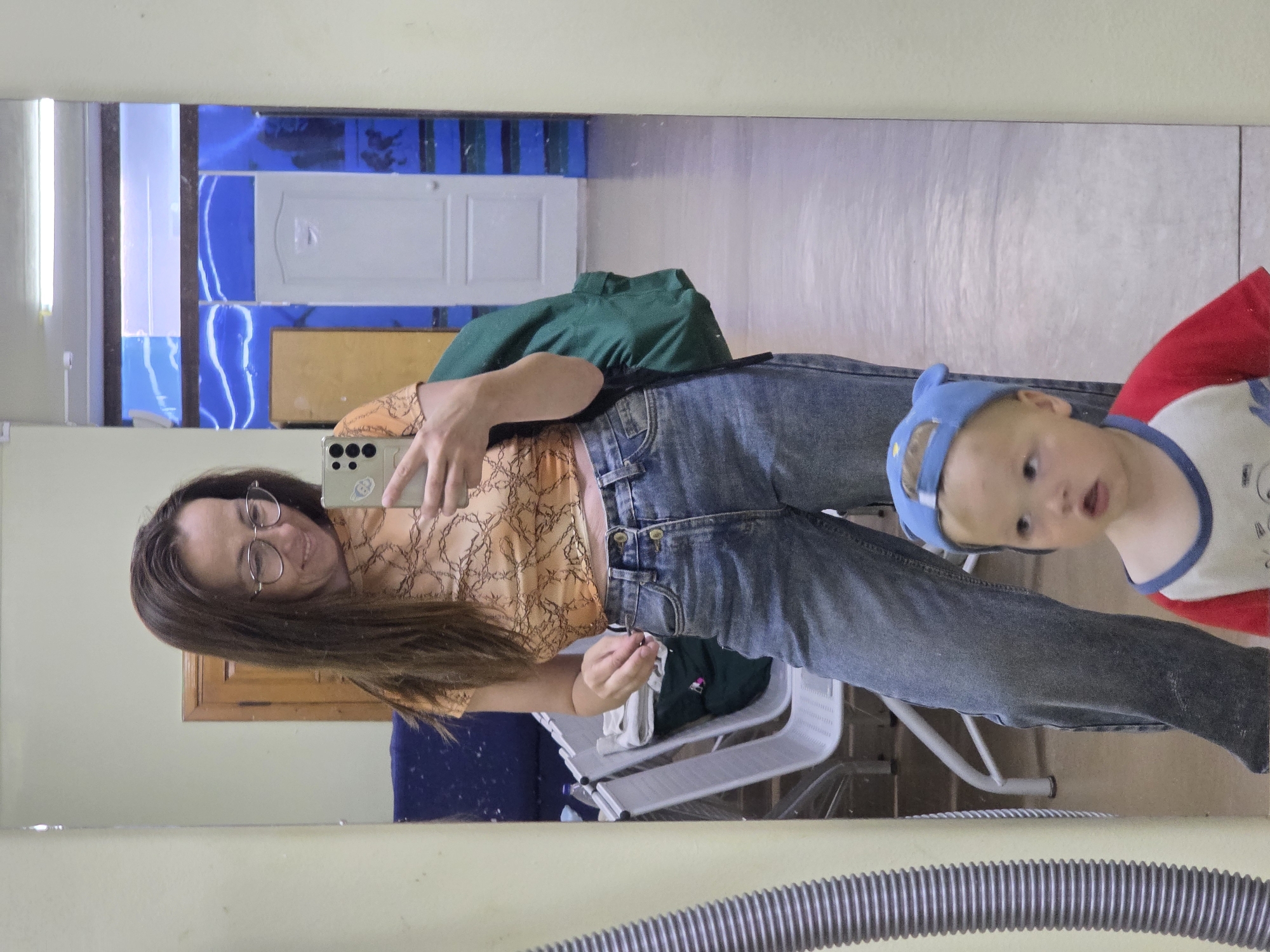 This is a mirror selfie taken by a woman who is wearing glasses, a light orange crop top, and blue jeans. She is holding a smartphone in her right hand, as if to take the picture. Her brown hair is long and falls over her shoulders. Next to her, also reflected in the mirror, is a young child wearing a blue cap and a red and white shirt. The child appears to be looking at the camera.
The background reflected in the mirror shows a room with blue and white tiled walls, and a wooden cabinet. There are chairs visible in the background. The overall setting appears to be indoors, possibly a room in a house or a public space. The lighting suggests it is daytime.
The woman's expression seems to be a slight smile, and the child has a curious expression. The picture is taken from a slightly angled perspective, likely due to the mirror.