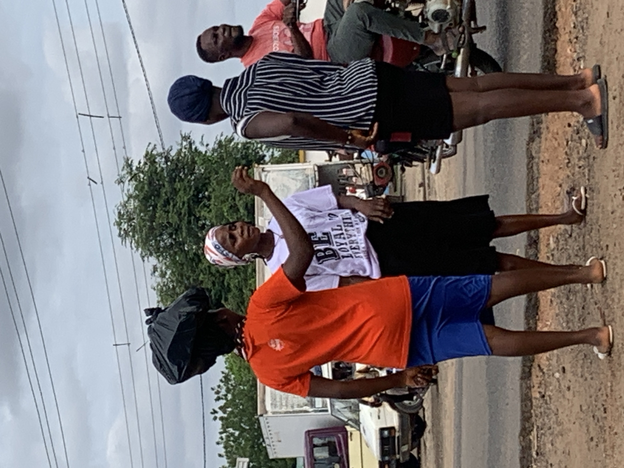 An outdoor scene in the city of Shendam, Nigeria, captures four individuals on an overcast day. In the foreground, a person with dark skin, wearing an orange t-shirt and blue shorts, stands facing right, balancing a black plastic bag on their head. They wear white flip-flops. To their right, an adult female with dark skin wears a white headwrap, a white t-shirt with partially visible text "BE LOYAL EVERYTHI," a black skirt, and white flip-flops. She stands facing left, gesturing with her right hand, index finger extended, towards the person with the bag on their head. Immediately to her right, a third person with dark skin stands with their back partially to the camera, wearing a dark blue headwrap, a black and white striped shirt, black shorts, and dark slide sandals. Further right, an adult male with dark skin is seated on a red motorcycle, facing right and slightly away from the other individuals. He wears a red t-shirt (with "COWBOYS" partially visible), dark trousers, and dark sandals. The ground beneath the individuals is unpaved dirt and gravel, transitioning to a paved road surface in the mid-ground. Various vehicles, including a white car and a white truck, are visible on the road and in the background. Green foliage and indistinct structures are present in the far background. Multiple power lines span across the cloudy, light grey sky.