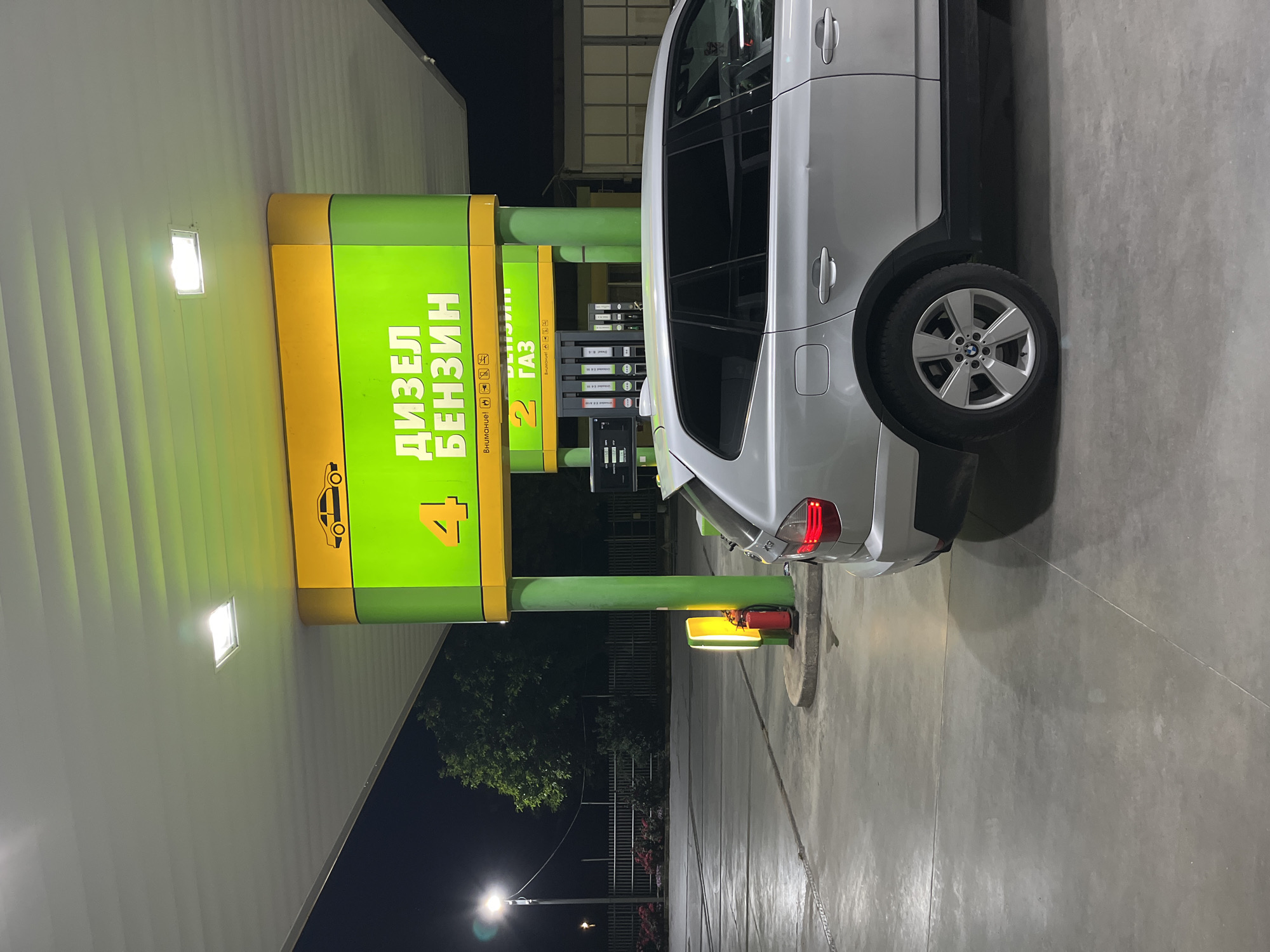 The image shows a silver BMW X3 parked at a gas station in Elhovo, Bulgaria. The gas station has yellow and green branding, with signs in Cyrillic script indicating the availability of diesel, gasoline, and gas. Pump number 4 is visible. The setting is at night, under artificial lighting. There are no people visible in the image. The car is positioned for fueling. A fire extinguisher is located near the pump. A streetlight and some trees can be seen in the background.