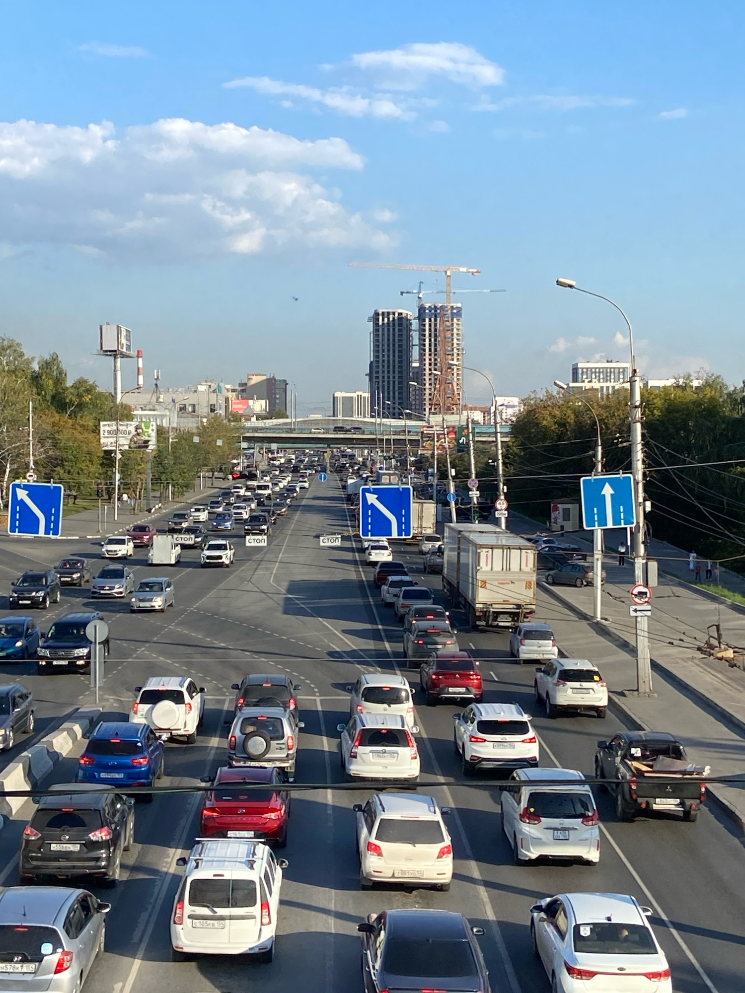 The image depicts a heavy traffic jam on a multi-lane road in Novosibirsk, Russia, under a clear blue sky with scattered clouds. The perspective is from an elevated position, looking down a long stretch of highway. Numerous cars, trucks, and vans are seen in various lanes, indicating slow-moving or stationary traffic. Road signs are visible, including blue directional signs with white arrows and "STOP" signs. The surrounding environment features trees with autumn foliage, several buildings in the background, and construction cranes, suggesting ongoing urban development. The overall scene captures a typical urban rush hour or congested period in the city.