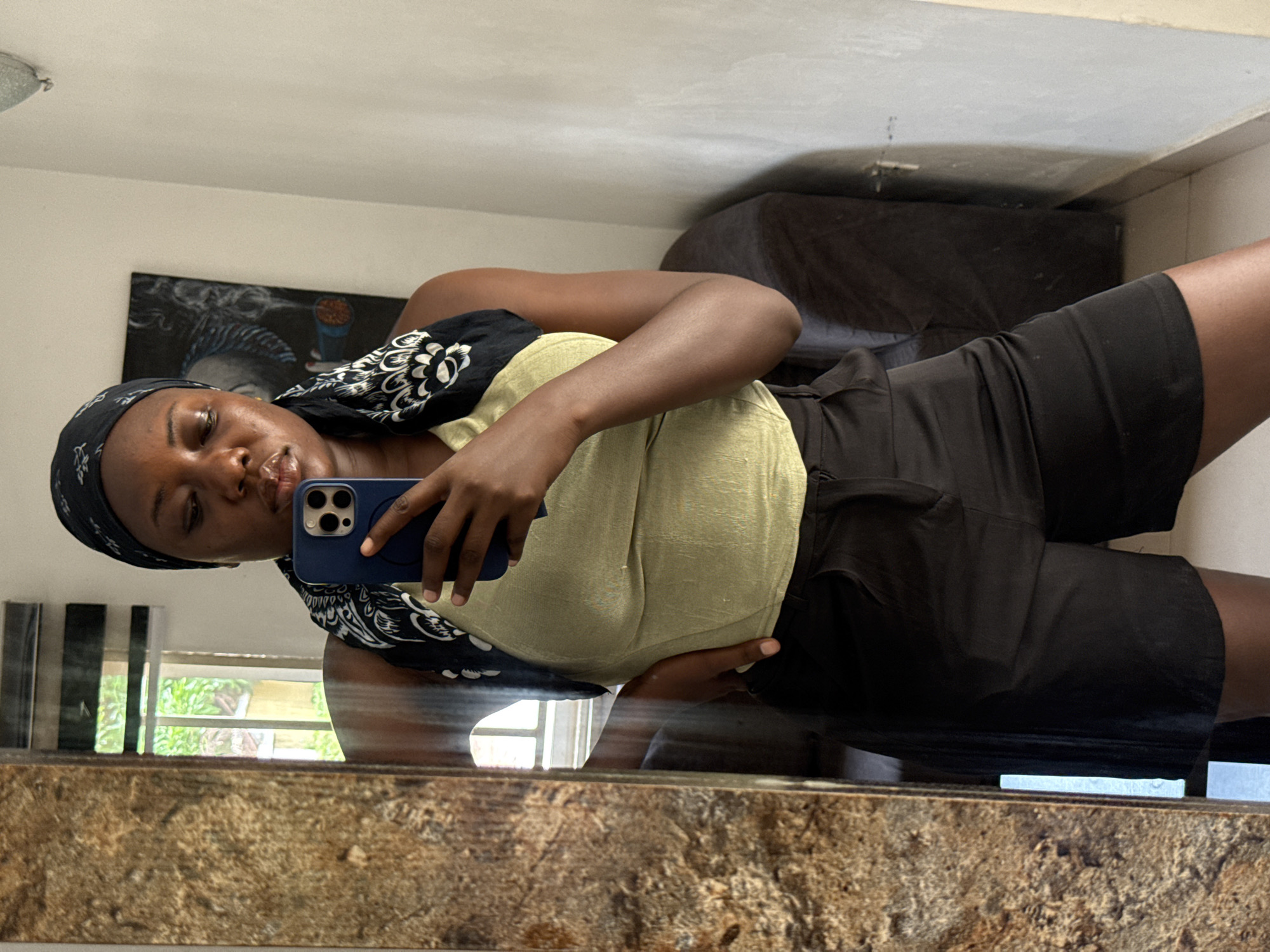 A dark-skinned woman is taking a selfie in what appears to be a residential setting. She is wearing a black headscarf with a white pattern, a light green top, and black shorts. She is holding a blue smartphone in her right hand. The photo is taken in front of a mirror with a brown, speckled frame. Behind her, there is a painting on the wall, a black couch, and a glimpse of the outdoors through a window with vertical blinds. The location is likely in a city, and based on the woman's ethnicity, it is plausible that it could be in Abuja, Nigeria.