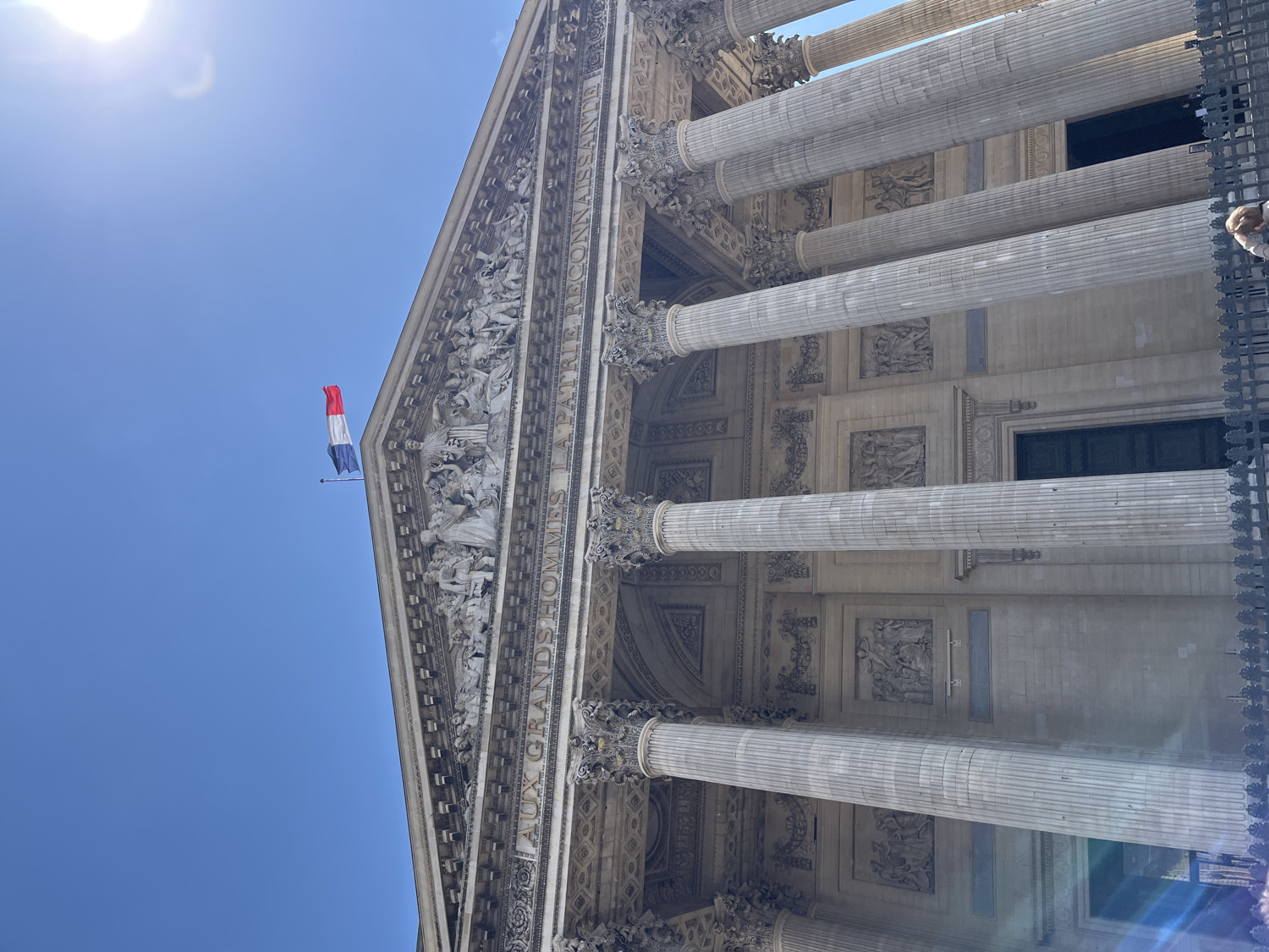 A low-angle shot captures the majestic facade of the Panthéon in Paris, France, under a clear, bright blue sky. The grand neoclassical building is seen from below, emphasizing its towering scale and classical architecture.
Dominating the upper section is a large pediment adorned with an elaborate sculptural group depicting numerous figures. Below the pediment, a prominent frieze bears the inscription "AUX GRANDS HOMMES LA PATRIE RECONNAISSANTE," which translates to "To the great men, the grateful homeland." Supporting this structure are multiple massive, fluted columns with ornate capitals, characteristic of the Corinthian order.
A French tricolour flag, with its blue, white, and red vertical stripes, flies from a flagpole positioned near the top of the pediment, gently unfurling against the sky. The bright sun creates a strong glare in the upper left of the frame, indicating midday or early afternoon on a sunny day.
Between the columns, the building's walls feature intricate relief carvings and rectangular, dark openings that appear to be windows or doorways. On the far right edge of the image, a dark, decorative metal fence is partially visible, and a small portion of a person's head can be seen behind it. The overall scene conveys a sense of historical grandeur and national pride.