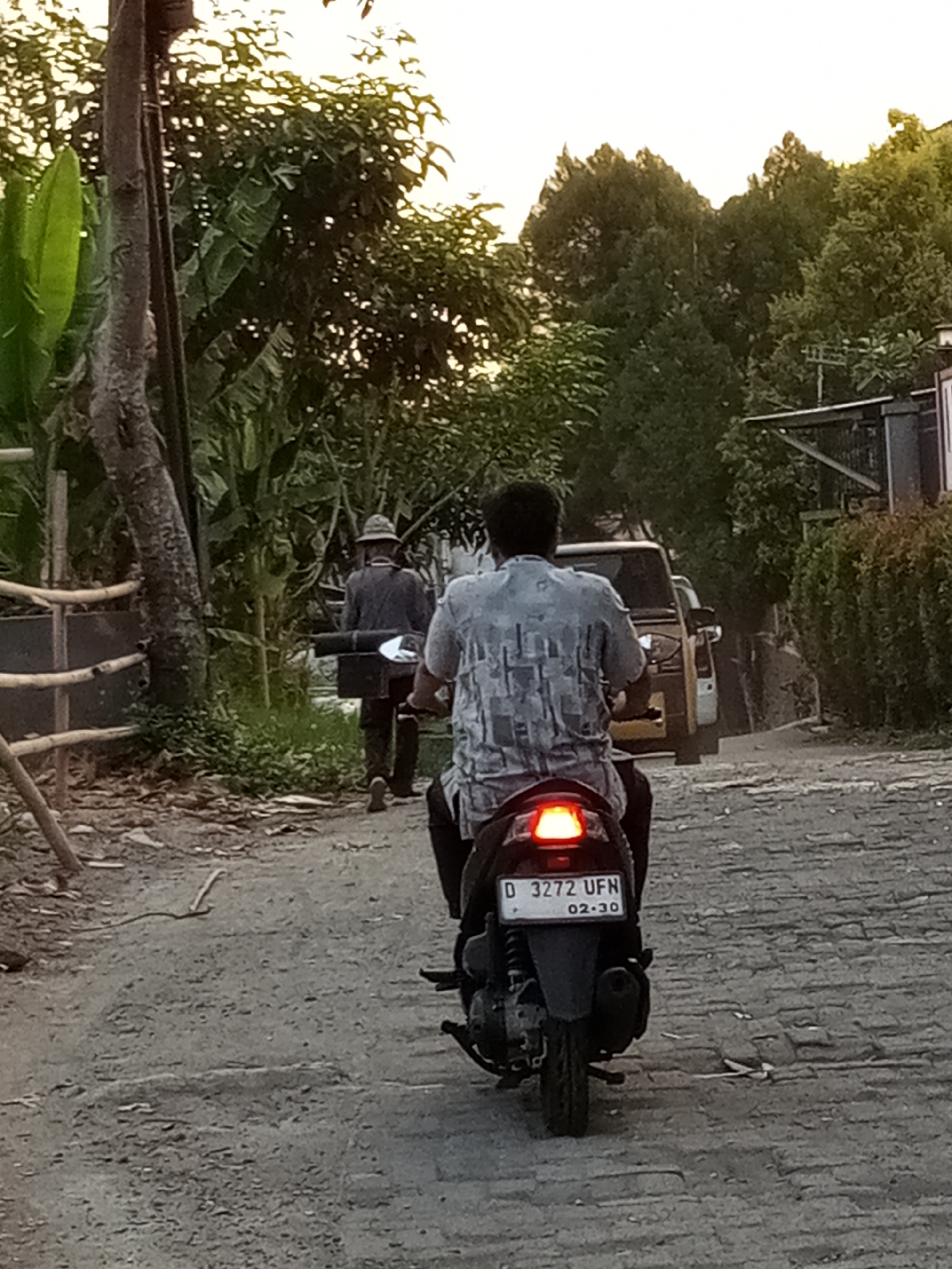 In the city of Cihanjuang, Indonesia, a man rides a motorcycle on a paved street. He is wearing a grey and white patterned shirt. His motorcycle has a license plate with the number "D 3272 UFN". Behind him, another man walks away from the camera, wearing a hat and dark clothing, carrying a black object. The street is lined with trees and vegetation on both sides, and a yellow vehicle is parked on the right side of the road. The scene appears to be set in the late afternoon or early evening.