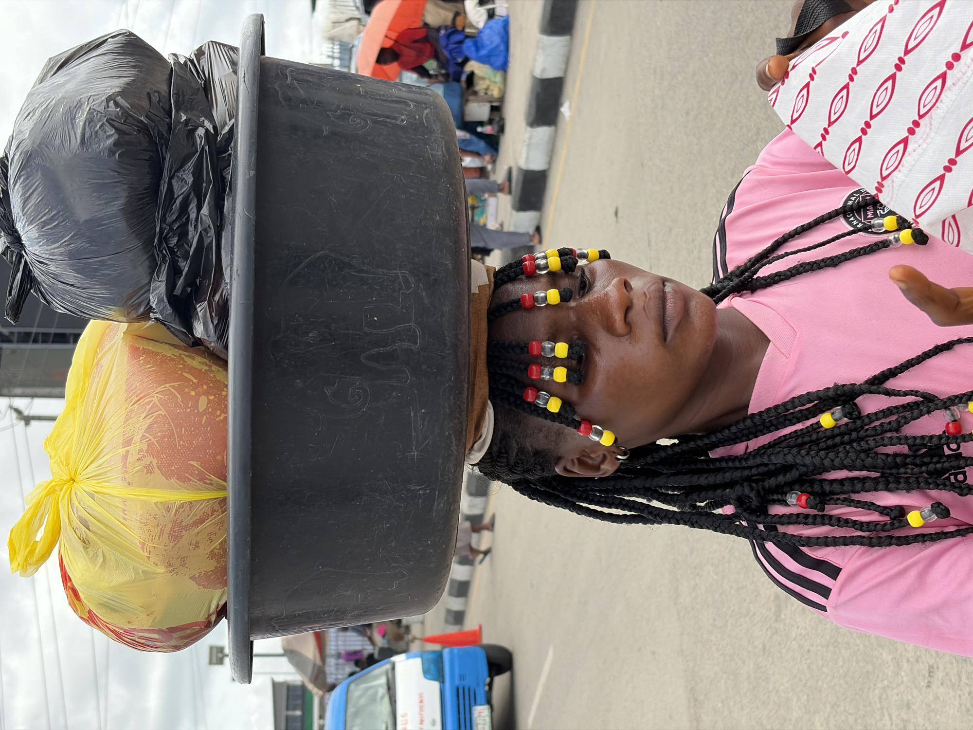 A person, presumably female, is carrying a large black plastic tub on their head, containing a large yellow bag at the bottom, and two black trash bags on top, walking on a paved street. The person is wearing a pink shirt with black stripes on the shoulder, and has braids with colorful beads in their hair. In the background there is a blue and white truck and many people milling around. The image was taken in Igboefon, Nigeria.