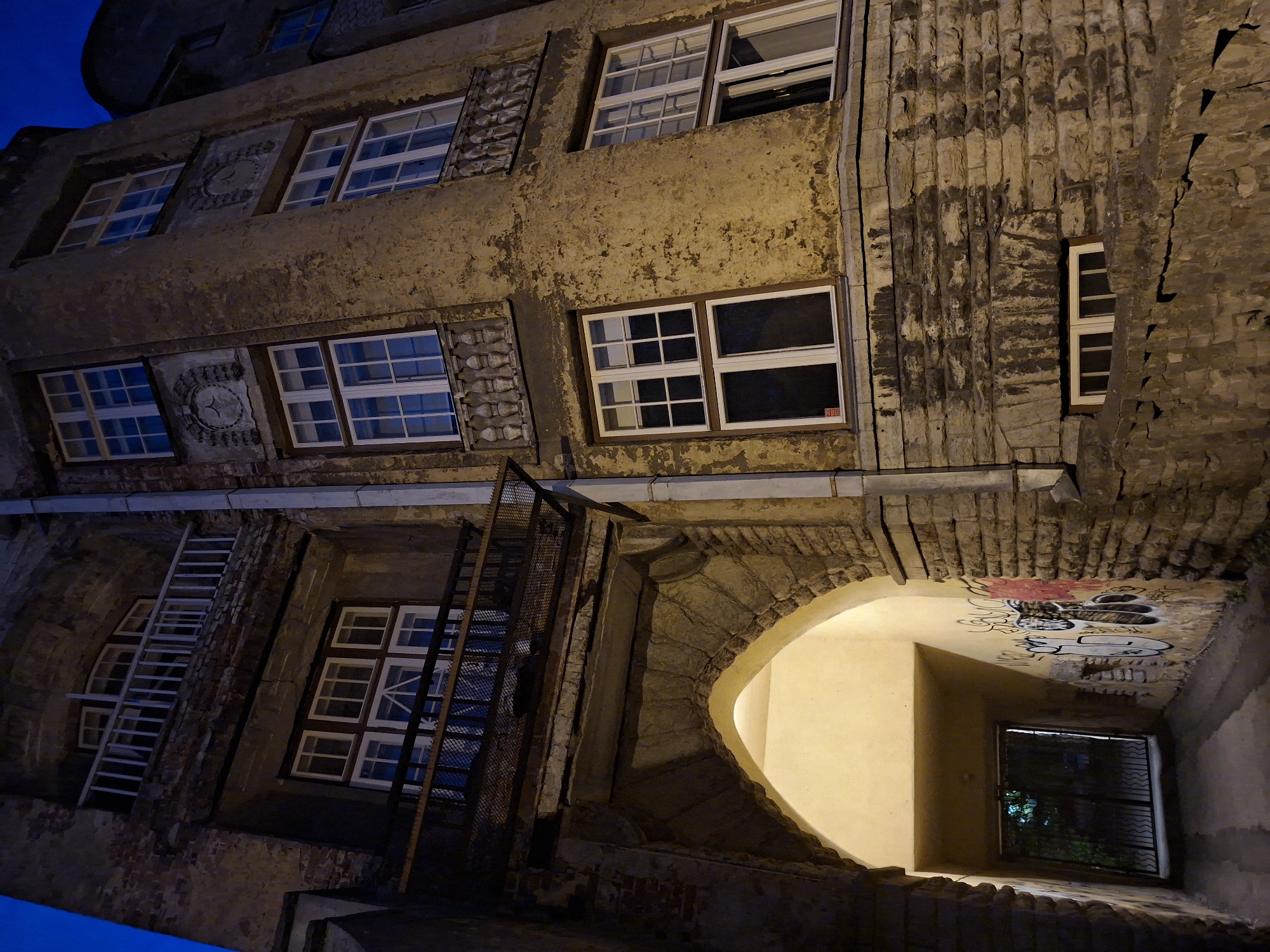 The image shows the exterior of an old, weathered building at night, located in Tallinn, Estonia. The building features a mix of brick and stucco, with peeling paint and visible signs of age. Several windows with white frames are visible, some with multiple panes. A balcony with a white railing is present on an upper floor, and a darker, metal-grated balcony is below it. An arched entryway leads into a dimly lit passageway, which has graffiti on its walls. A metal gate is visible at the end of the passageway. The overall scene conveys a sense of historical architecture in an urban setting at dusk or night.