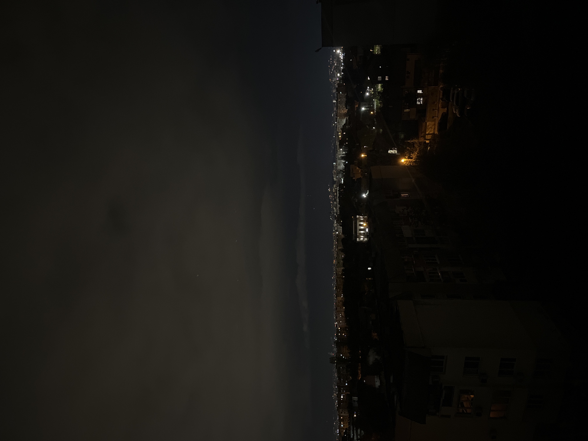 A nighttime cityscape is captured, presumably from a high vantage point, like a balcony or rooftop. The city, heavily implied to be Sofia, Bulgaria due to the building style, is illuminated with streetlights and building lights. A significant portion of the frame is filled with a dark sky, potentially obscured by clouds or smog. The right side of the image appears darker, possibly due to the presence of closer buildings blocking the light. Specific landmarks are not visible, but the general layout of the city with its varied lighting is discernible. The image is taken at a vertical orientation.