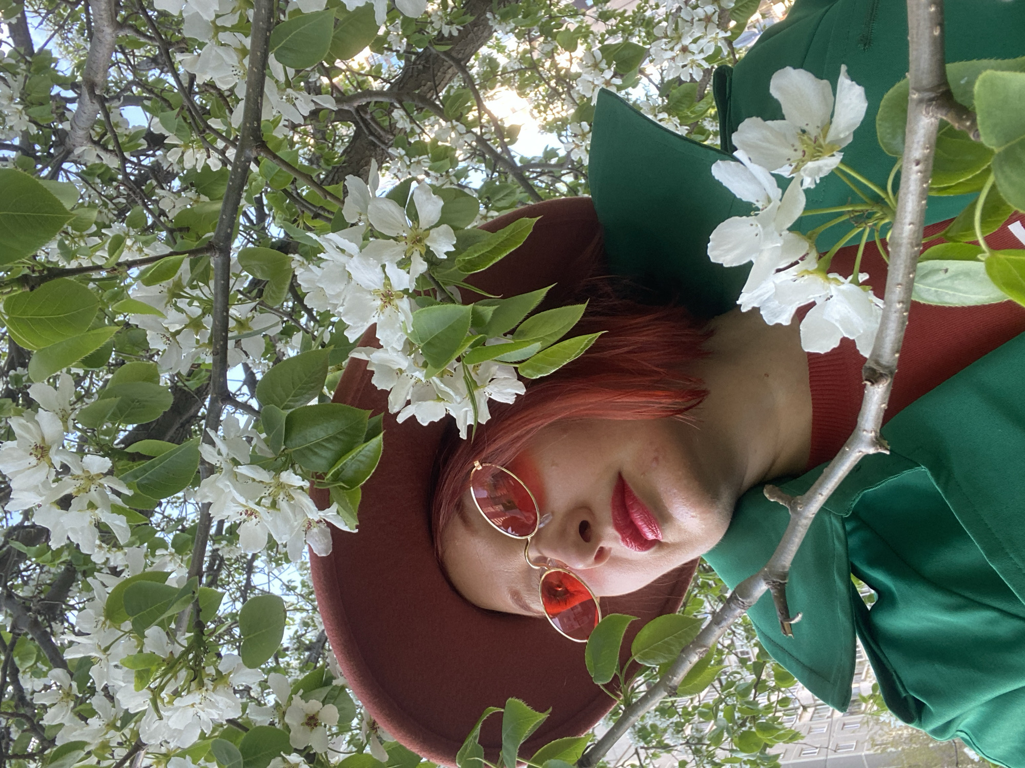 A person is shown in an upward-facing shot. The individual has red hair and is wearing red lipstick and round, red-tinted sunglasses. They are wearing a rust-colored hat and a green collared jacket with a red undershirt. The person is surrounded by branches with white flowers and green leaves, partially obscuring a tall building in the background. The picture was taken in Yekaterinburg, Russia.