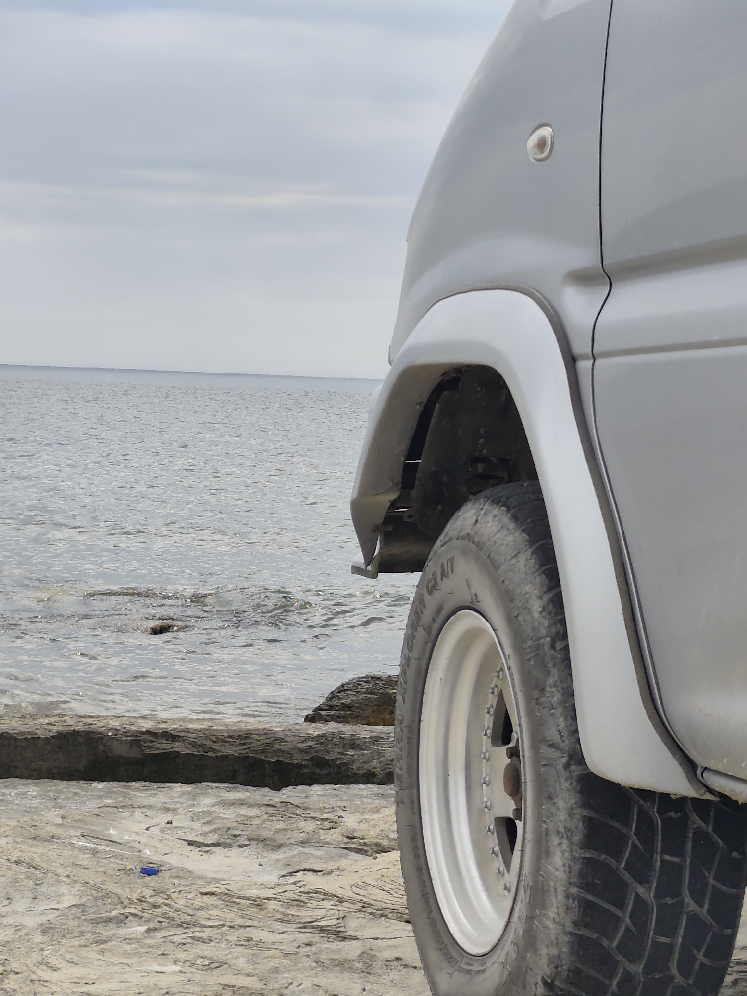 This is a medium shot, eye-level, outdoor photograph of a silver van parked on a rocky shore by the Caspian Sea in Aqshuqyr, Kazakhstan. The van is positioned in the right half of the frame, with its front wheel and fender visible. The wheel is a polished alloy rim with a black tire. The van's bodywork is a smooth, light gray color, and a small amber turn signal is visible on the side. The ground beneath the van is a mixture of sand and rough, layered rocks. In the background, the calm, grey waters of the Caspian Sea extend to the horizon, meeting a pale, overcast sky. The water is slightly choppy in places where it meets the rocks. A few scattered rocks are visible in the water.