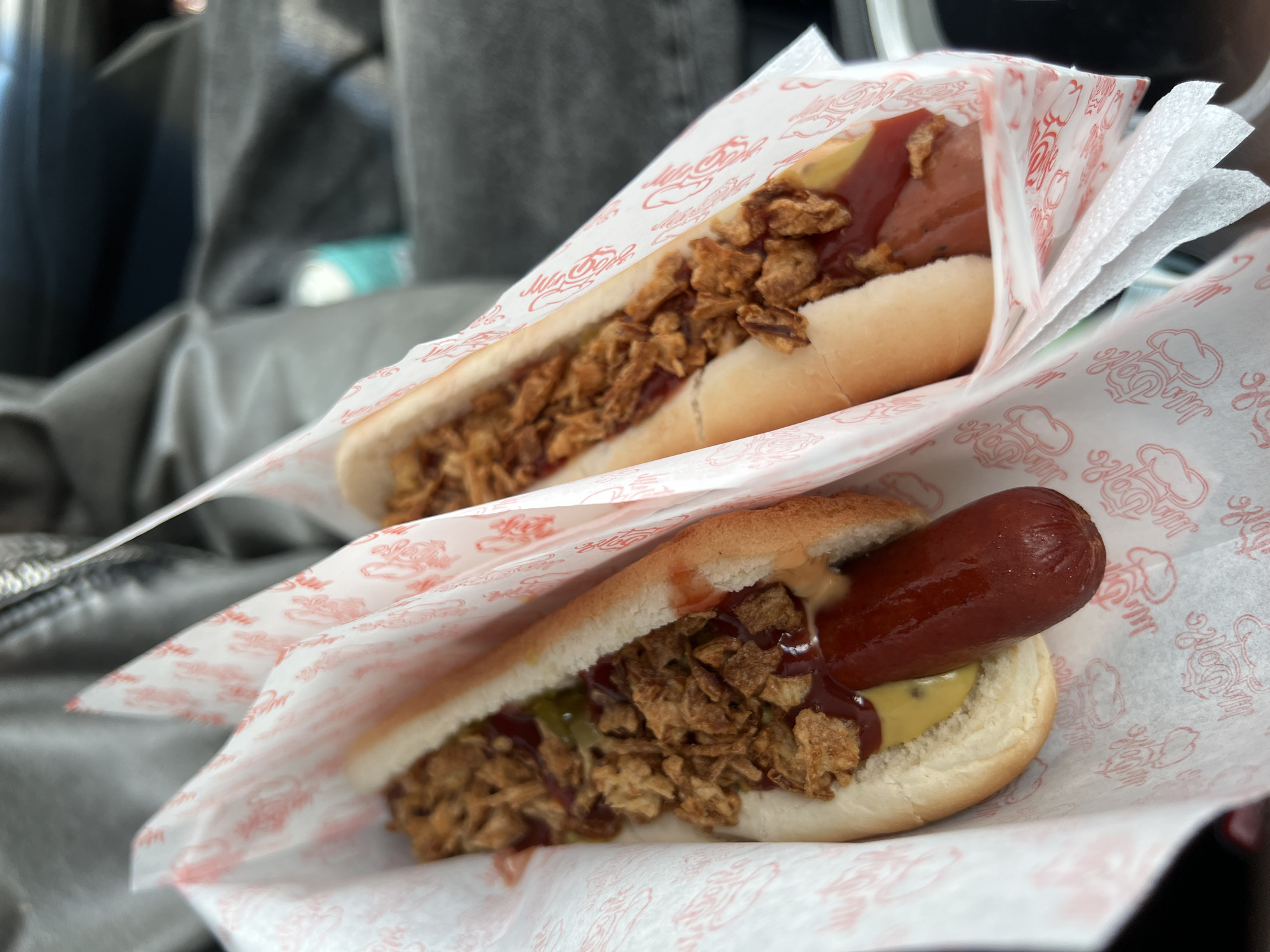 Two hot dogs are presented in white paper wrappers patterned with red script. Each hot dog sits in a bun and is topped with ketchup, mustard, and fried onions. The image suggests a casual setting, potentially a car or takeaway situation. The location details (city and Unknown) are unknown.