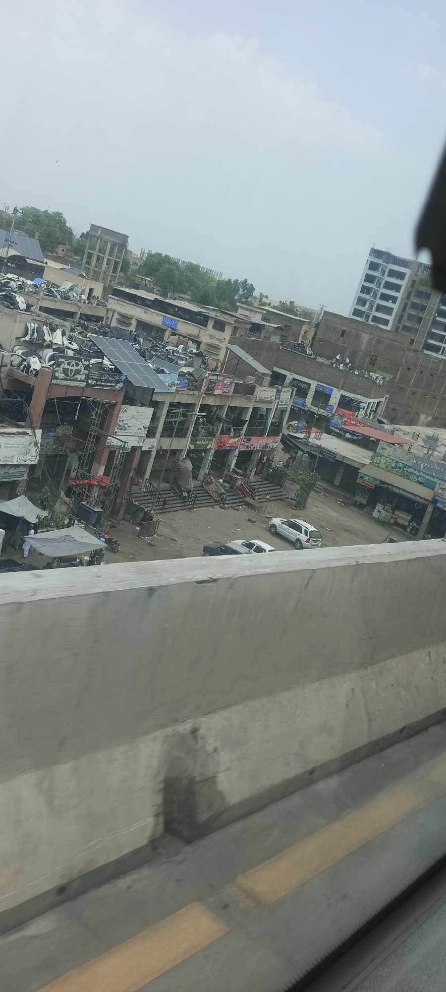 The image shows an aerial view of a commercial area in Alam Khan Kalay, Pakistan, likely a market for automotive parts and services. In the foreground, a concrete barrier runs horizontally across the bottom of the frame, obscuring the lower portion of the scene.
The main subject is a multi-story building complex with a weathered and somewhat dilapidated appearance. The buildings are a mix of concrete and brick structures, some with open storefronts and exposed rebar, suggesting ongoing construction or disrepair. The roofs of several buildings are covered with solar panels, indicating an effort towards renewable energy.
The buildings are densely packed with what appear to be automotive parts, including car bodies, bumpers, and various machinery, piled high on rooftops and in front of shops. Several vehicles are visible in the street and within the complex: a white SUV, a white sedan, and a black sedan are parked. A motorcycle is also present.
There are a few individuals visible. Two men in white clothing are standing under a grey canopy near the bottom left of the building complex, possibly vendors or customers. Another person, also in white, is seen near a motorcycle. A few other figures are indistinctly visible on the balconies and rooftops of the buildings.
The environment appears to be an urban or semi-urban setting. In the background, more buildings and trees are visible, along with a large water tower. The sky is overcast. The overall impression is of a busy, functional marketplace with a focus on the automotive industry.