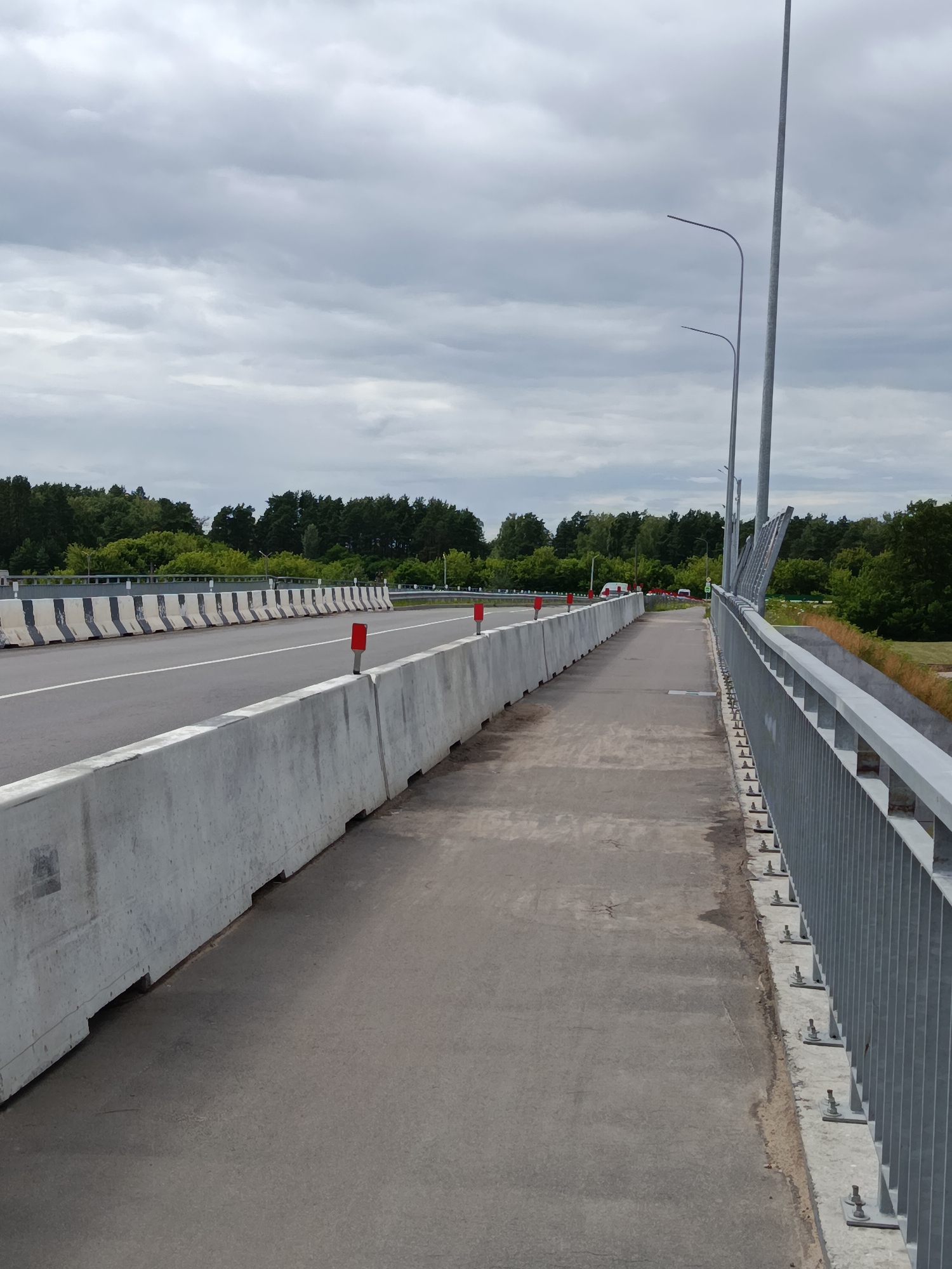 The image captures a view of a bridge in Svietlahorsk, Belarus, on a cloudy day. The bridge has a pedestrian walkway separated from the road by a concrete barrier. The road itself is bordered by a striped concrete barrier. Streetlights are visible on the right side of the image, and trees line the horizon in the background. A few cars are present on the road, and red markers are placed along the concrete barriers.