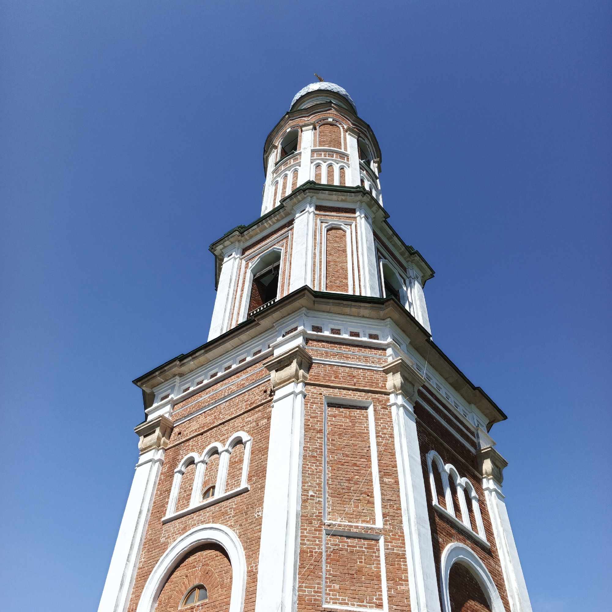 The image shows the bell tower of the Church of the Intercession of the Most Holy Theotokos in Tugustemir, Russia. The bell tower is constructed of red brick with white decorative accents and topped with a grey dome. The sky is a clear, vibrant blue. The perspective is a low-angle shot, looking up at the tower against the sky. There are no people or other objects visible in the frame.