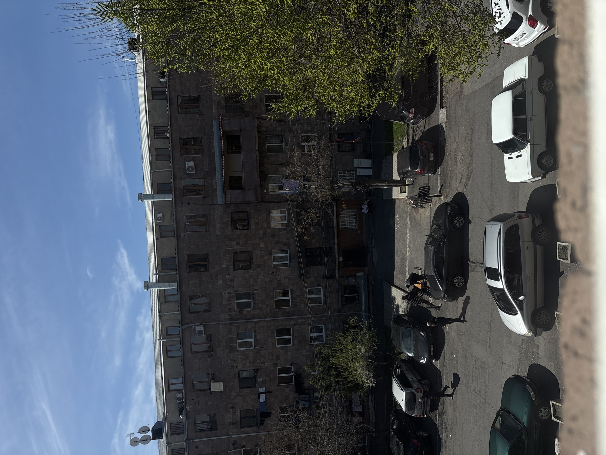 The image shows a courtyard in Yerevan, Armenia, lined with a multi-story residential building on the left. The building is constructed with dark stone, has a flat roof, and features multiple windows and air conditioning units mounted on the facade. A blue sky with sparse clouds is visible above the building.
In the courtyard, several parked cars are arranged along the side of the building. People are walking or standing amongst the vehicles. There is a small tree near the center of the image, partially obscuring some of the parked cars. Other trees with green foliage are visible near the building.
