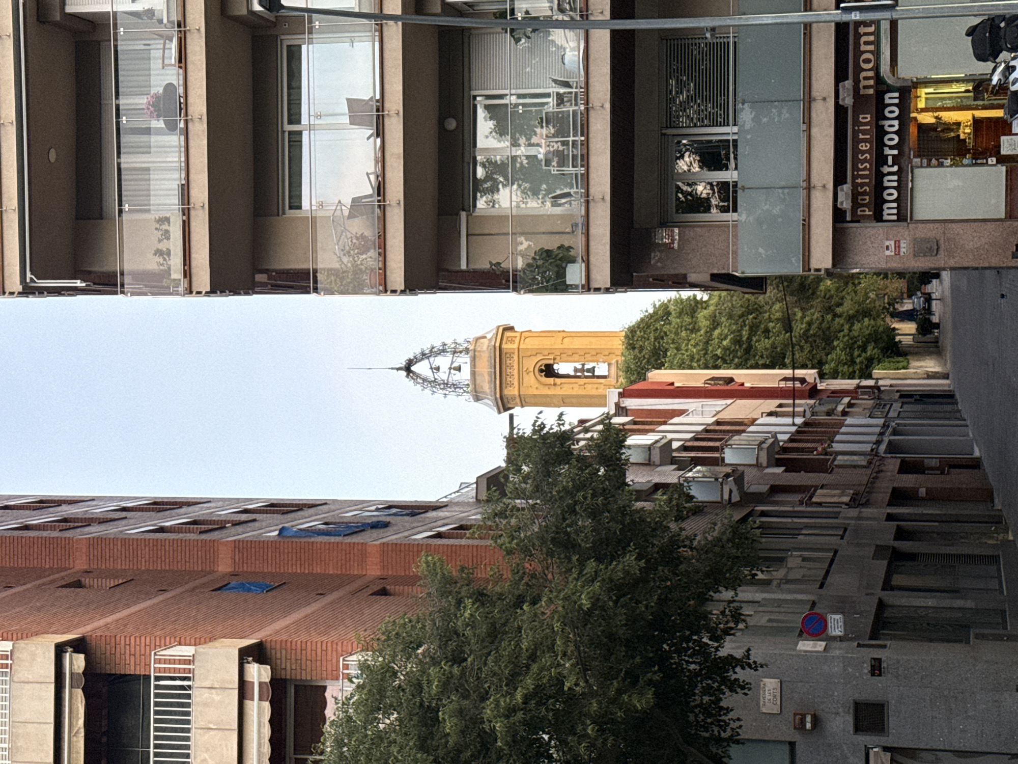 The image shows a narrow street in Barcelona, Spain, lined with buildings. At the end of the street, a yellow church tower is visible, framed by the buildings on either side. The buildings are of varying heights and architectural styles, with balconies, windows, and signage. A large tree is growing in the foreground, partially obscuring some of the buildings. The image is oriented with the top of the frame pointing to the left. A sign for "pastisseria mont-rodon" is visible on one of the buildings. The sky is overcast. There is a motorbike with two riders in the distance, and other figures walking on the sidewalk. There is a traffic sign in the lower right corner.