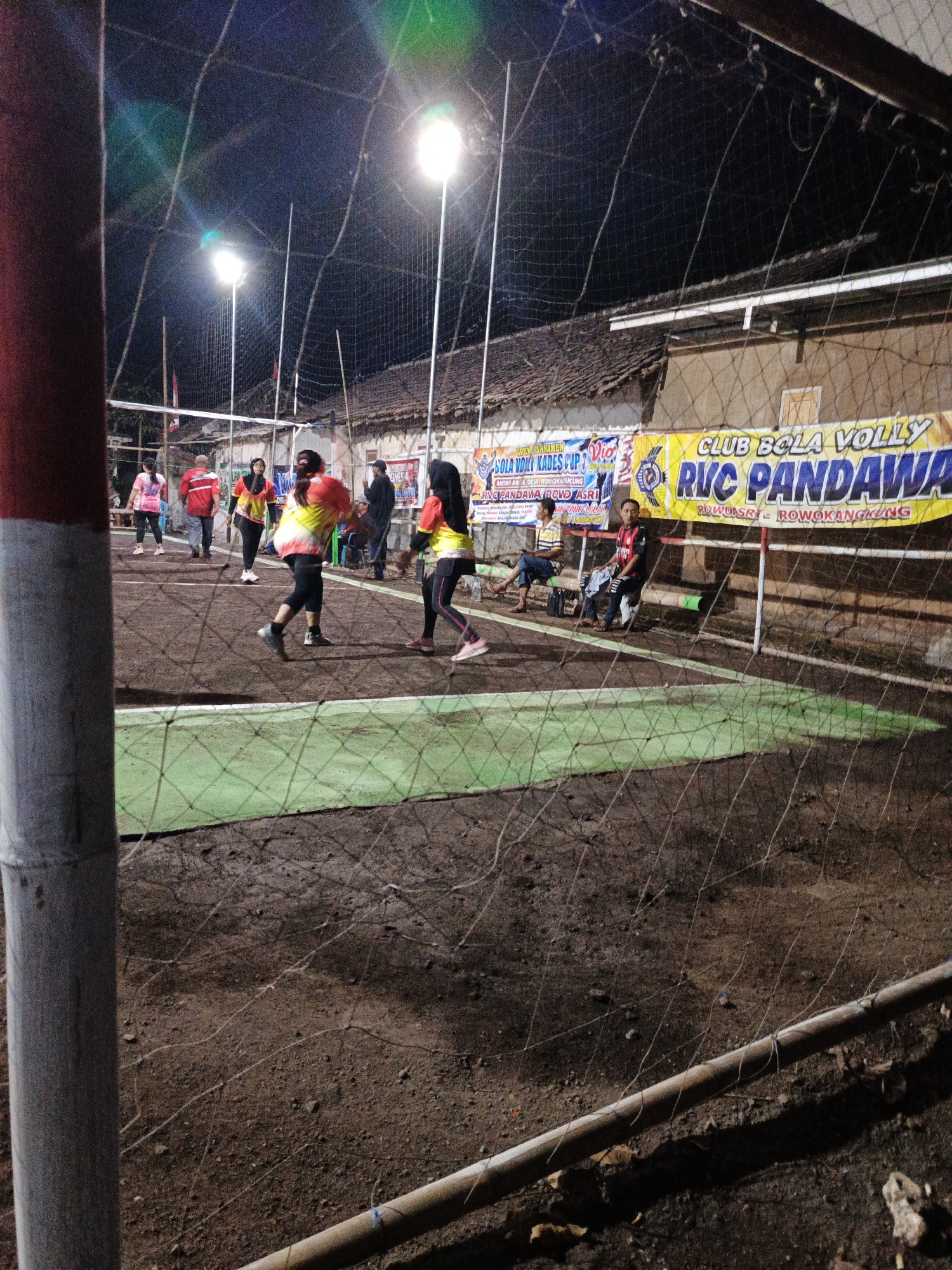 This is a photograph of an outdoor volleyball game at night in Rowokangkung, Indonesia. The court appears to be made of dirt with a green boundary line. A net divides the court, and a volleyball pole stands on the left side of the frame. Bright floodlights illuminate the court, casting shadows.
There are several people visible on the court, engaged in play. On the left side of the net, three individuals are positioned: one person in a pink shirt and dark pants, another in a red shirt and dark pants, and a third in a yellow and orange shirt with dark pants, facing away from the camera. On the right side of the net, two people are actively playing: one in a yellow and orange shirt with dark pants, in mid-action, and another in a similar colored shirt and black leggings, also in motion.
In the background, behind the net, several spectators are seated on benches. One man in a yellow striped shirt sits on the left, and another man in a red and black shirt is seated in the center. Further back, a banner with Indonesian text and logos is displayed on a building. The building's roof is visible, made of clay tiles. The overall environment suggests a local community sporting event taking place after dark.