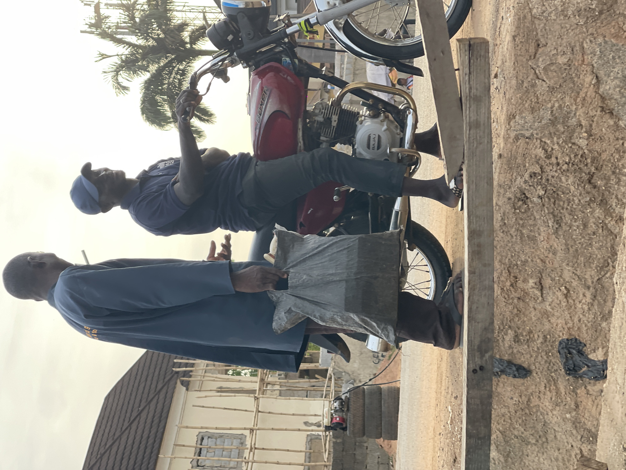 This image, captured in Abuja, Nigeria, depicts an outdoor scene featuring two men and a motorcycle, likely in the late afternoon or early evening given the soft, warm lighting.
One man is seated on a red motorcycle, oriented sideways, wearing a blue polo shirt, dark trousers, and a blue cap. He is holding a smartphone in his right hand, extended as if taking a photo or video, or perhaps viewing its screen. His feet are bare. The motorcycle's fuel tank clearly displays "BOXER," and the engine block shows "BAJAJ."
Standing next to the motorcycle, facing the first man, is another man dressed in a dark blue, long-sleeved garment. Some yellow text is visible on the back of his shirt, but it is too indistinct to read clearly. He is holding a dark, rectangular package, possibly wrapped in plastic, with both hands, positioned between himself and the motorcycle. He wears dark sandals.
The background reveals a developing area with a partially constructed building featuring bamboo scaffolding. A palm tree is visible in the upper portion of the frame. The ground is rough, appearing to be a mix of dirt and concrete, with loose wooden planks scattered nearby. The overall scene suggests a casual interaction or transaction taking place on a street or roadside.