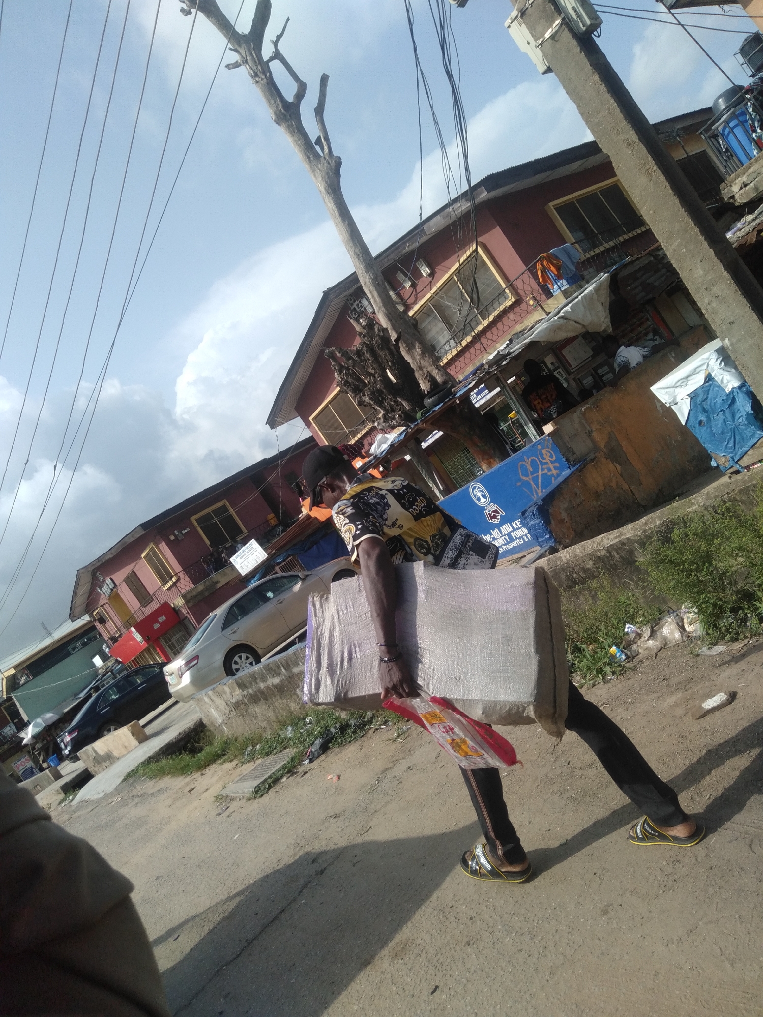 A person carries a large object in an urban environment. The person wears dark trousers, sandals, and a patterned shirt, with a hat or cap visible. The object they are carrying is large and rectangular, possibly fabric. They also carry a red and white bag in their other hand. The scene is a street in a city, with buildings lining the roadside and parked cars in the background. There is a utility pole and wires overhead. The sky is cloudy. The location is the city of Unknown, Unknown.