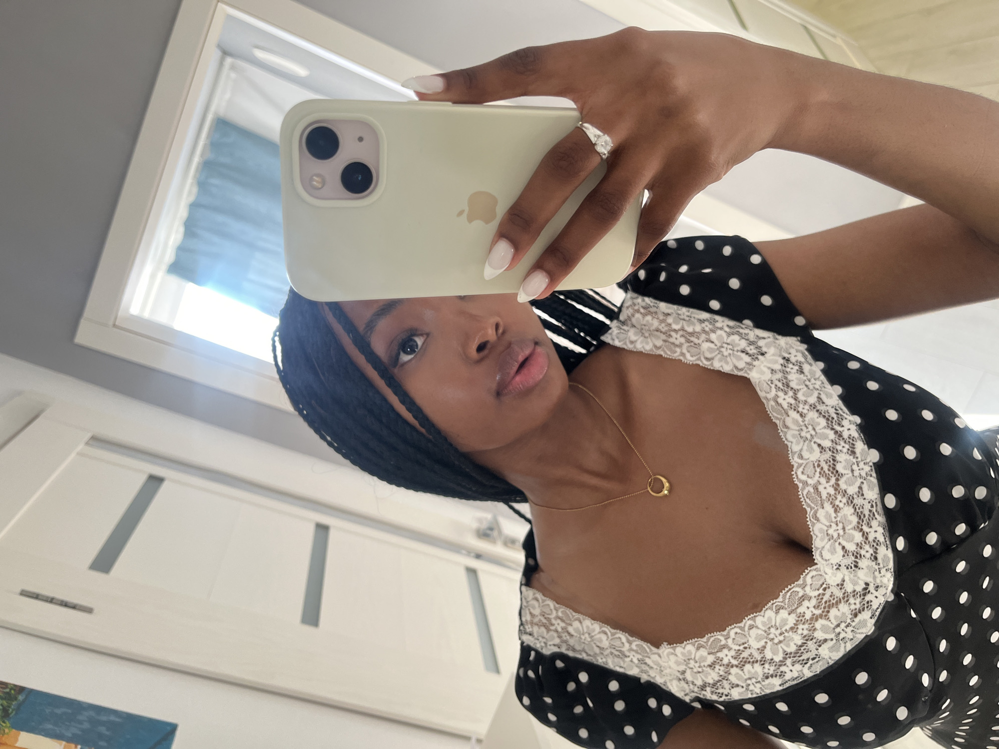 A young Black woman with braided hair takes a mirror selfie in a room. She is wearing a black and white polka dot dress with lace trim. She holds a light green smartphone in her right hand, which is also adorned with a silver ring. The background shows a white door with grey inserts and a reflection of a window with blue curtains. The overall impression is of a casual, indoor setting, likely a home or hotel room. There is no information or visual cue to suggest this location is in Kazan, Russia.