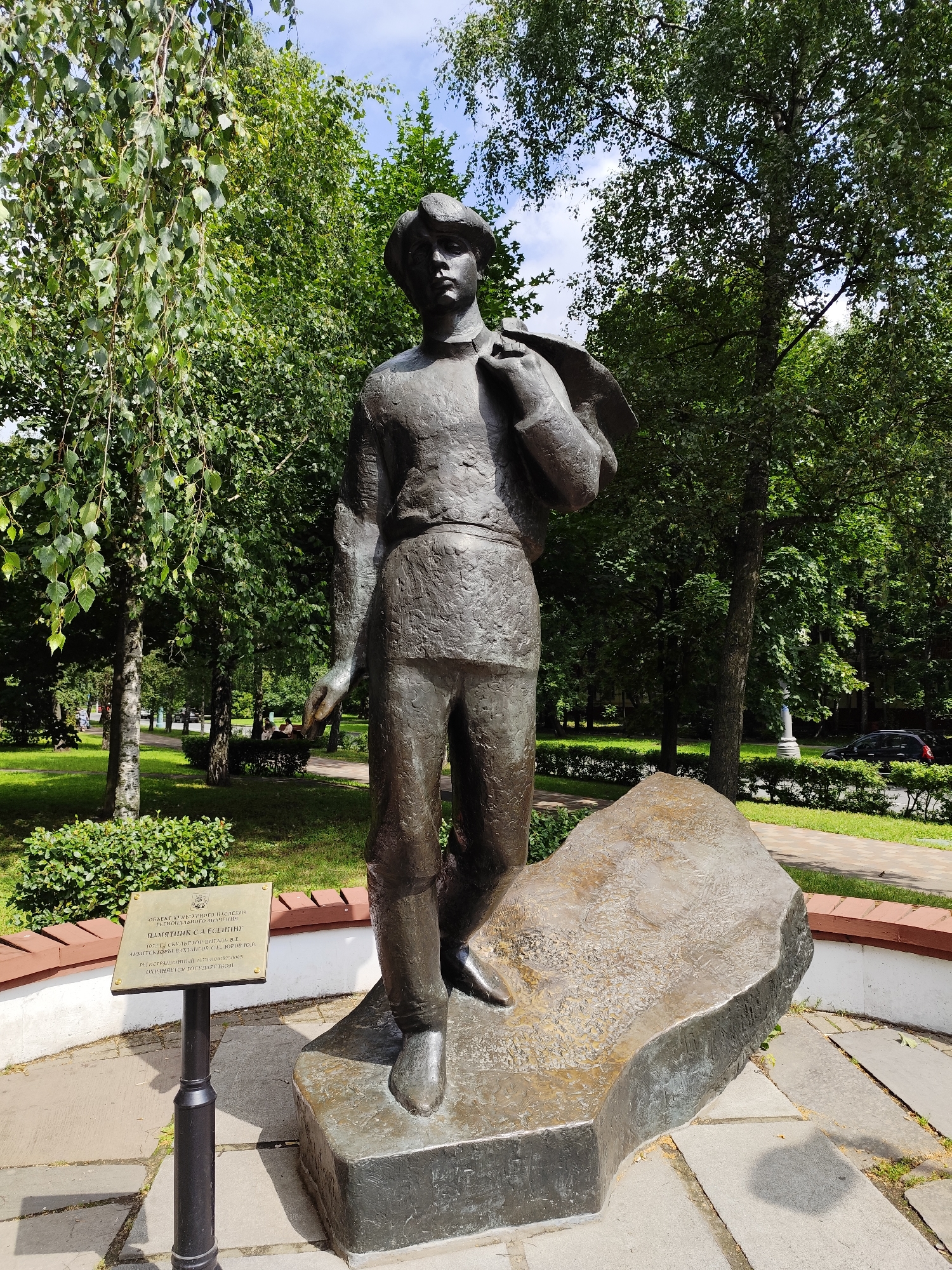 A full-body bronze statue of a young man wearing a cap and carrying a jacket over his shoulder stands on a rough, uneven rock base. He is depicted in a walking pose, facing forward and slightly to the viewer's left. The statue is located outdoors in a park-like setting with lush green trees and a blue sky with white clouds visible in the background. A paved walkway and a low, red-capped brick wall are present in the foreground. A small plaque on a black metal stand is positioned to the left of the statue. In the distance, several people are seated on benches and a car is visible on a road. This statue is located in Moscow, Russia, specifically at the corner of Bolshaya Nikitskaya Street and Gnezdnikovsky Lane.