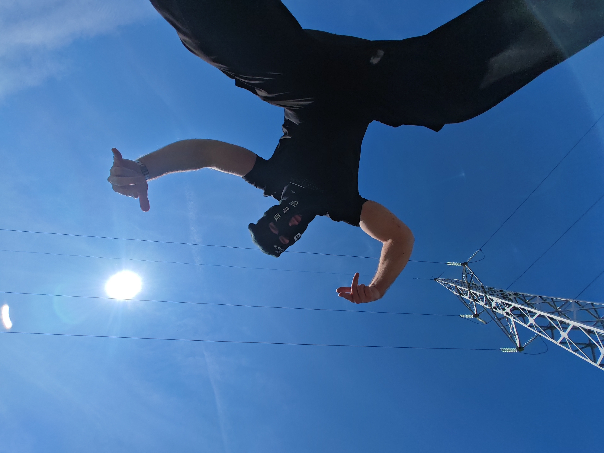 The image displays a low-angle perspective looking upwards towards a bright, clear blue sky with minimal cloud cover. A single individual is positioned in the upper half of the frame, appearing inverted, with their torso and legs visible. The person is wearing black pants, a black short-sleeved shirt, and a black balaclava covering their face, featuring light-colored text or symbols. Their right arm is extended to the left, with the hand forming a "thumbs up" gesture and a striped wristband visible. Their left arm is also extended, with the hand open and fingers slightly curled. In the upper left quadrant, the sun is visible as a bright, star-like burst, creating lens flare. Multiple thin, dark power lines horizontally traverse the sky, intersecting the sun and the individual. In the lower right quadrant, a large, metallic lattice-work electricity transmission pylon, light in color, extends diagonally upwards, featuring multiple green-tinged ceramic insulators. The scene is consistent with an outdoor location featuring high-voltage power lines and a transmission pylon, characteristic of industrial or peripheral zones within or around Shumen, Bulgaria.