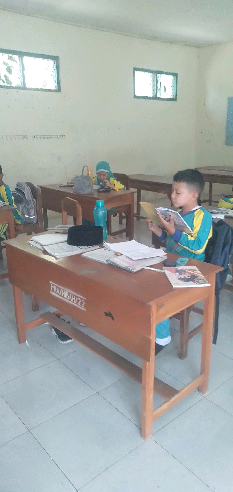 A classroom setting in Plemahan, Indonesia, features two young students engaged in learning. On the right, a boy wearing a blue and yellow long-sleeved shirt and turquoise pants sits at a wooden desk, intently reading a book. On the left, another child, wearing a yellow shirt and a light green headscarf, also sits at a desk, appearing to focus on an object on the desk. Several wooden desks are arranged in rows, with some school bags and books visible on them. A blue water bottle sits on one of the desks. The room has light-colored walls and a tiled floor. The overall impression is one of quiet concentration and academic activity.