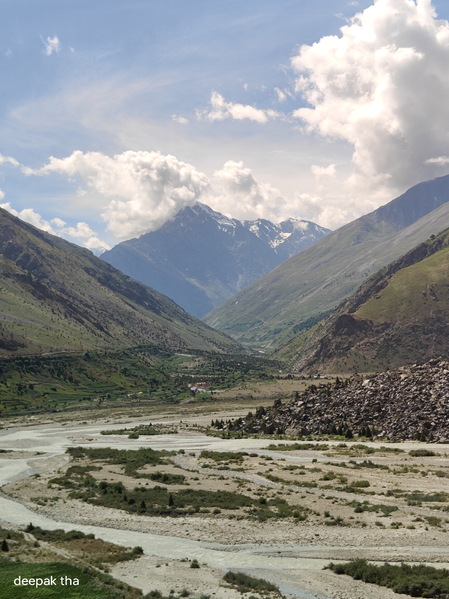 The image depicts a wide, arid valley in Darcha, India, characterized by a winding river flowing through a bed of sand and stones. Steep, rocky mountains flank the valley on both sides, with patches of green vegetation clinging to their lower slopes. The sky is a bright blue, dotted with fluffy white clouds. In the foreground, the river braids into multiple channels, separated by sandy islands and sparse scrub. The middle ground shows more vegetation, including terraced fields and scattered trees, leading towards a small cluster of buildings nestled in the valley. The background is dominated by towering, snow-capped mountains that disappear into the clouds. A watermark "deepak tha" is visible in the bottom left corner.