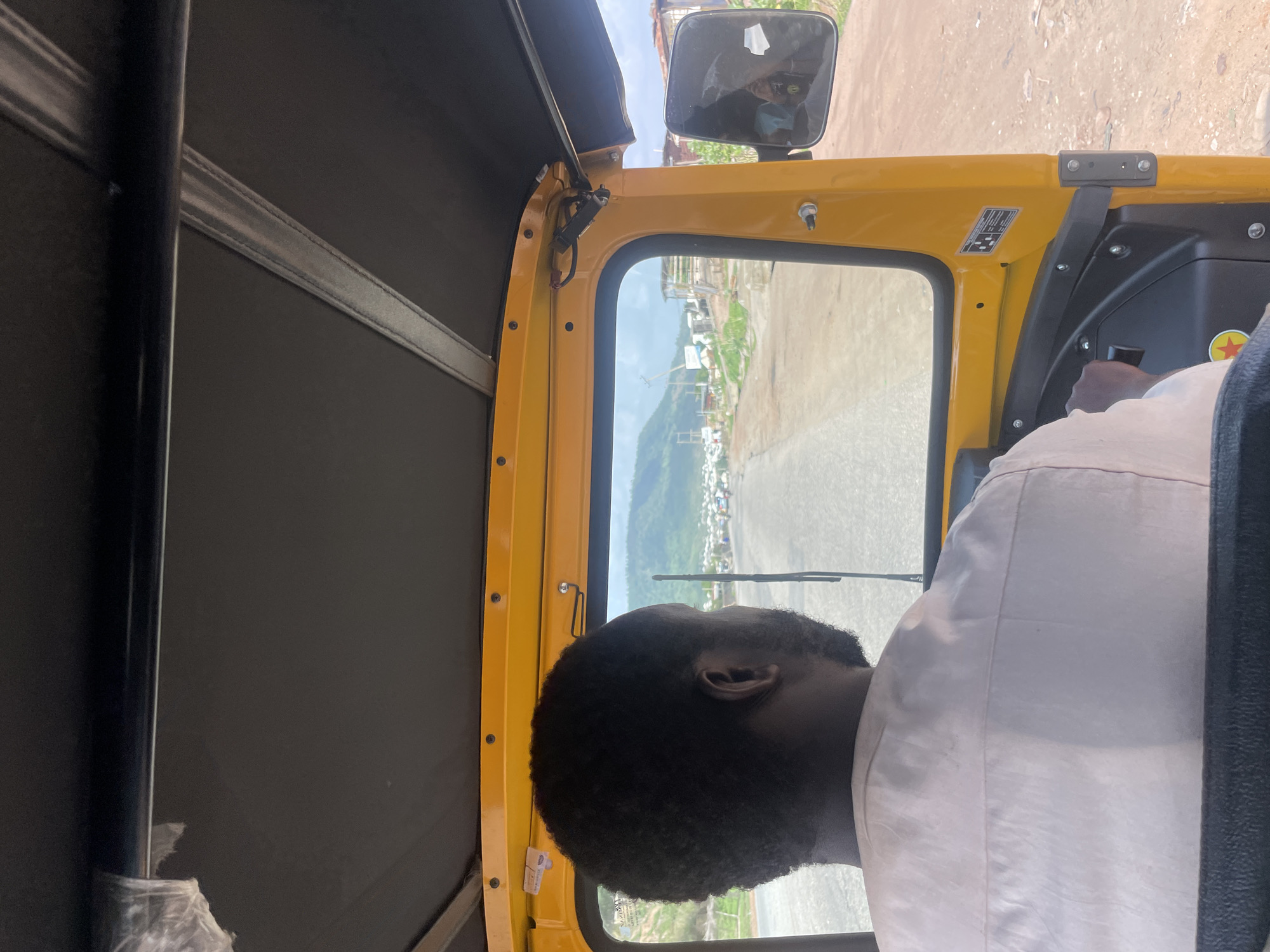 The image captures a view from inside a yellow vehicle, possibly an auto-rickshaw or a similar three-wheeled vehicle, in what is interpreted as the city of Lokoja, Nigeria. A black man with dark hair and a beard is visible, wearing a white shirt. He is seated behind the wheel, seemingly in the driver's seat. The vehicle's window frames a scene of a road leading into a town or city. The landscape in the background includes rolling green hills, likely marking the outskirts of the city. The vehicle's side mirror is visible in the top right. The scene suggests movement, implying the vehicle may be in transit.