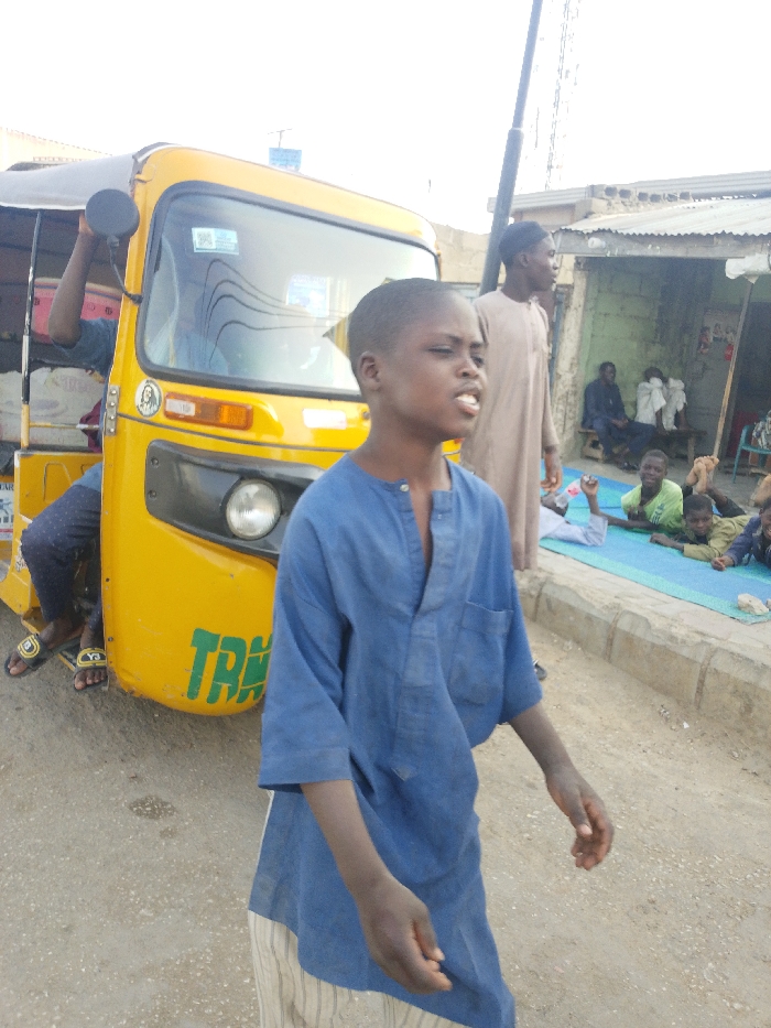 The image displays a daytime street scene in Garun Gawa, Nigeria, featuring multiple individuals and a yellow three-wheeled auto-rickshaw. In the foreground, a young boy with short dark hair, wearing a blue long-sleeved tunic and light-striped trousers, walks towards the right with his mouth slightly open. To his left, a bright yellow auto-rickshaw, commonly known as a Keke Napep, is visible, marked with green "TRA" lettering on its front lower panel and a sticker of a figure (possibly Jesus) on the windshield. A person's leg, clad in dark patterned trousers and black flip-flops with yellow "Y-3" branding, extends from the left side of the rickshaw, while another arm holds a black object inside. Behind the foreground boy, a young man in a light brown traditional long garment and matching cap stands, looking rightward. In the background, on a raised concrete sidewalk, several children are reclined or lying on a blue mat, including one child in a light green shirt smiling towards the camera. Further back, two adult males are seated on a wooden bench in front of a simple building with concrete walls and a corrugated metal roof. The road surface is a light gray asphalt or packed dirt, and a utility pole is visible on the right side of the frame under a bright, hazy sky.