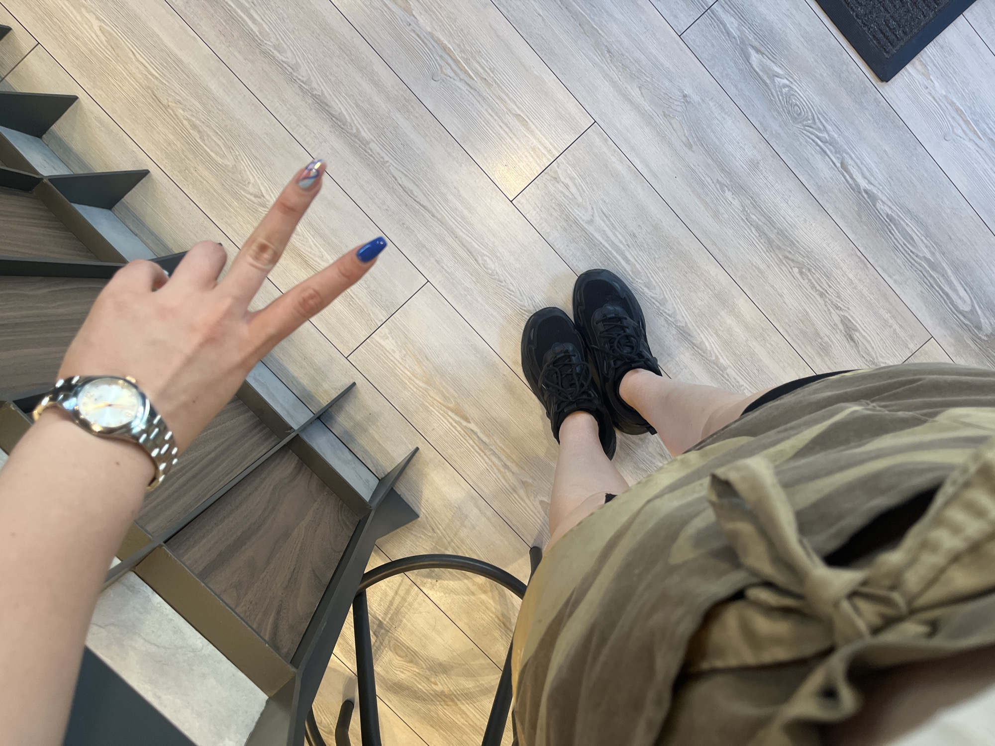 The image shows a top-down perspective of a person's legs and arm. The person is wearing black athletic shoes and a camouflage-patterned garment that appears to be a skirt or dress. Their left arm is visible, wearing a silver wristwatch. The hand is making a peace sign with two fingers extended. The background consists of a light-colored wooden floor and part of a dark-colored structure with metallic accents. The lighting suggests an indoor environment. The location is not specified in the image.