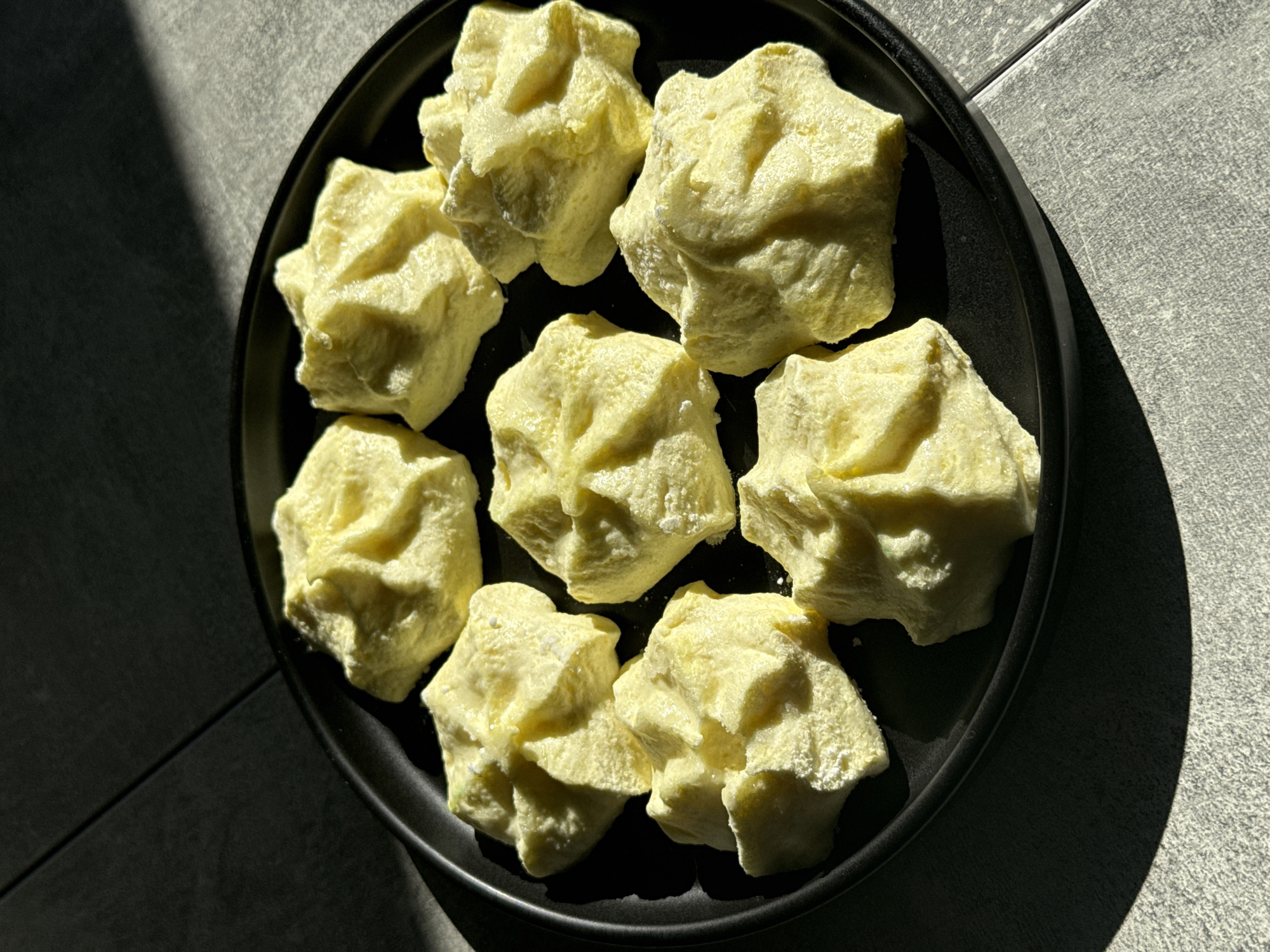 The image displays eight pale yellow, irregularly shaped solid pieces, possibly meringues, cookies, or another type of food, arranged on a round, matte black plate. Each piece exhibits a textured, somewhat crumbly or porous surface, suggesting a delicate or sugary consistency. They are scattered rather than neatly arranged on the plate.
The scene is illuminated by strong, directional natural light, likely sunlight, casting sharp, elongated shadows onto the plate and the surrounding surface. These shadows create a high-contrast visual, highlighting the contours of the objects and the rim of the plate.
The background features a light grey tiled surface, indicating a floor or countertop, with clear grout lines forming a grid pattern. Part of the tiled area is brightly lit by the sunlight, while another significant portion is in deep shadow, emphasizing the strong light source. The overall aesthetic is minimalist, focusing on the texture and form of the pieces and the interplay of light and shadow. There is no visible text in the image. The setting is an indoor environment, likely during the daytime, in Tomilino, Russia.
