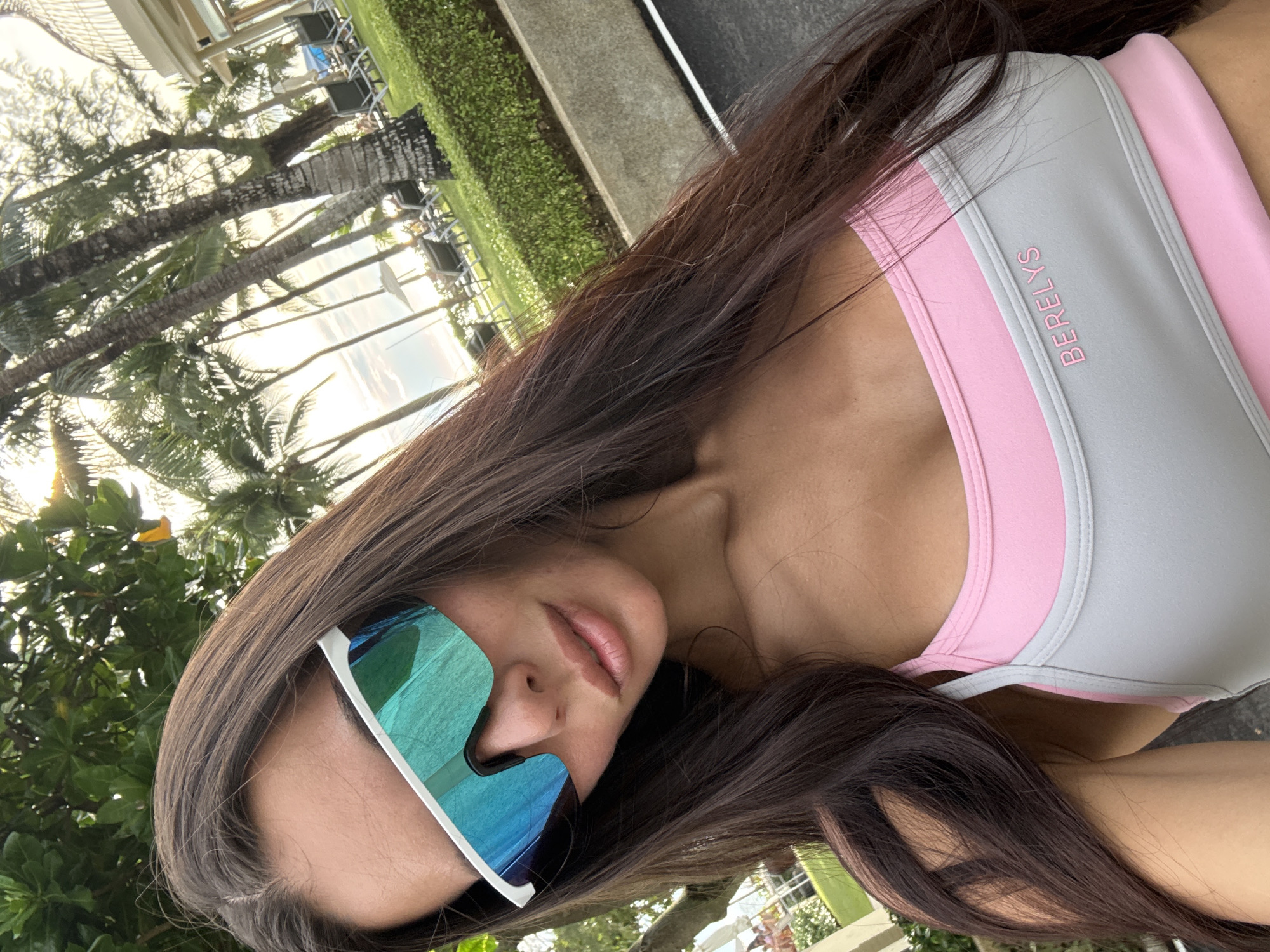 A woman with long, dark hair, wearing large, white-framed sunglasses with reflective blue and green lenses, and a pink and gray sports bra with the word "BERELYS" embossed on the front, poses for a selfie outdoors. The background suggests a tropical resort in Karon, Thailand, with palm trees, lush green bushes, lounge chairs, and glimpses of buildings and the sky. The lighting indicates it is daytime, possibly late afternoon, with the sun casting a warm glow. The woman is the central focus of the image.